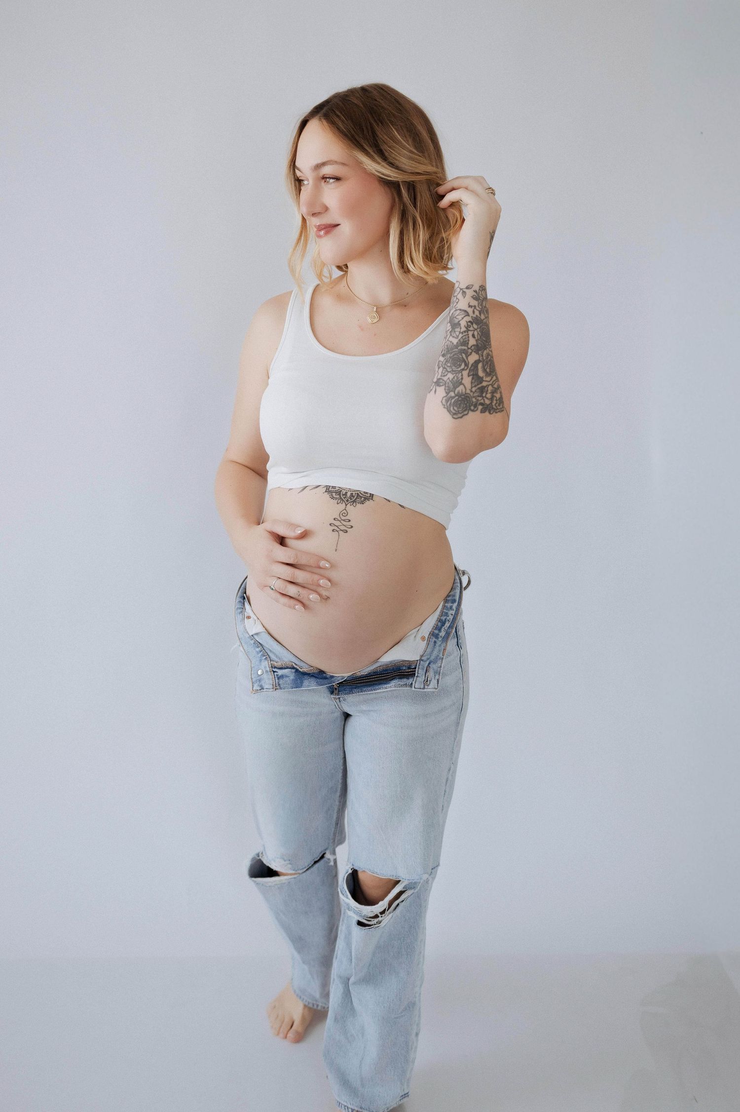 Casual maternity portrait showing expecting mother in distressed jeans and white top against light backdrop.