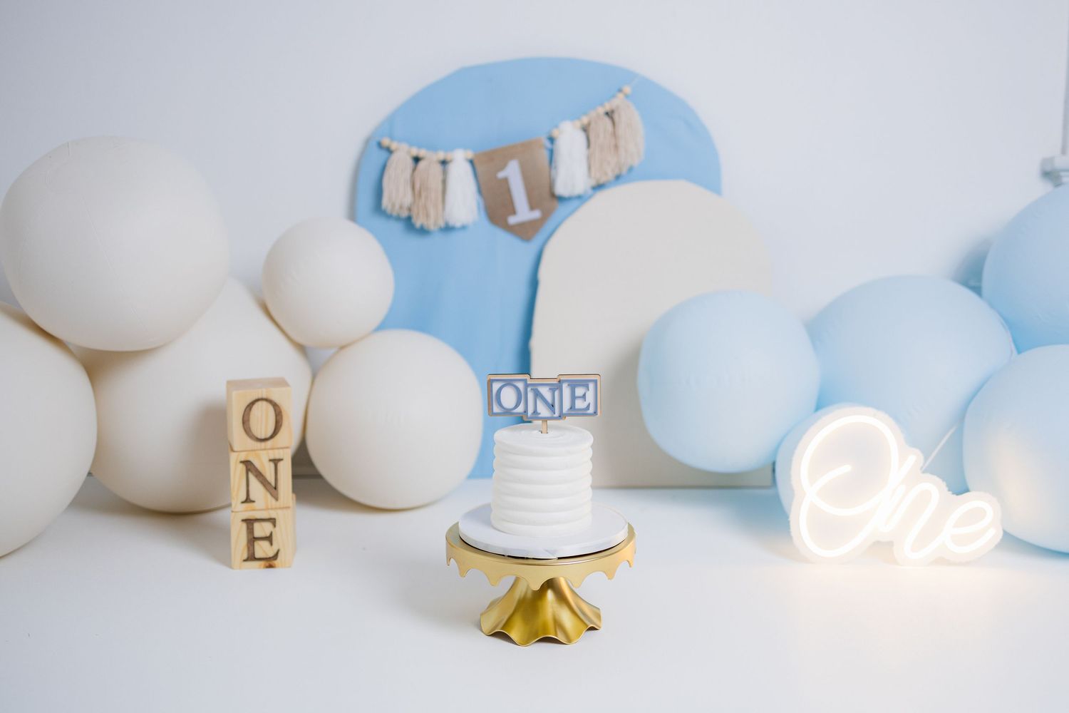 Minimalist baby boy first birthday setup with blue balloons, wooden blocks spelling 'ONE', and a small cake stand.