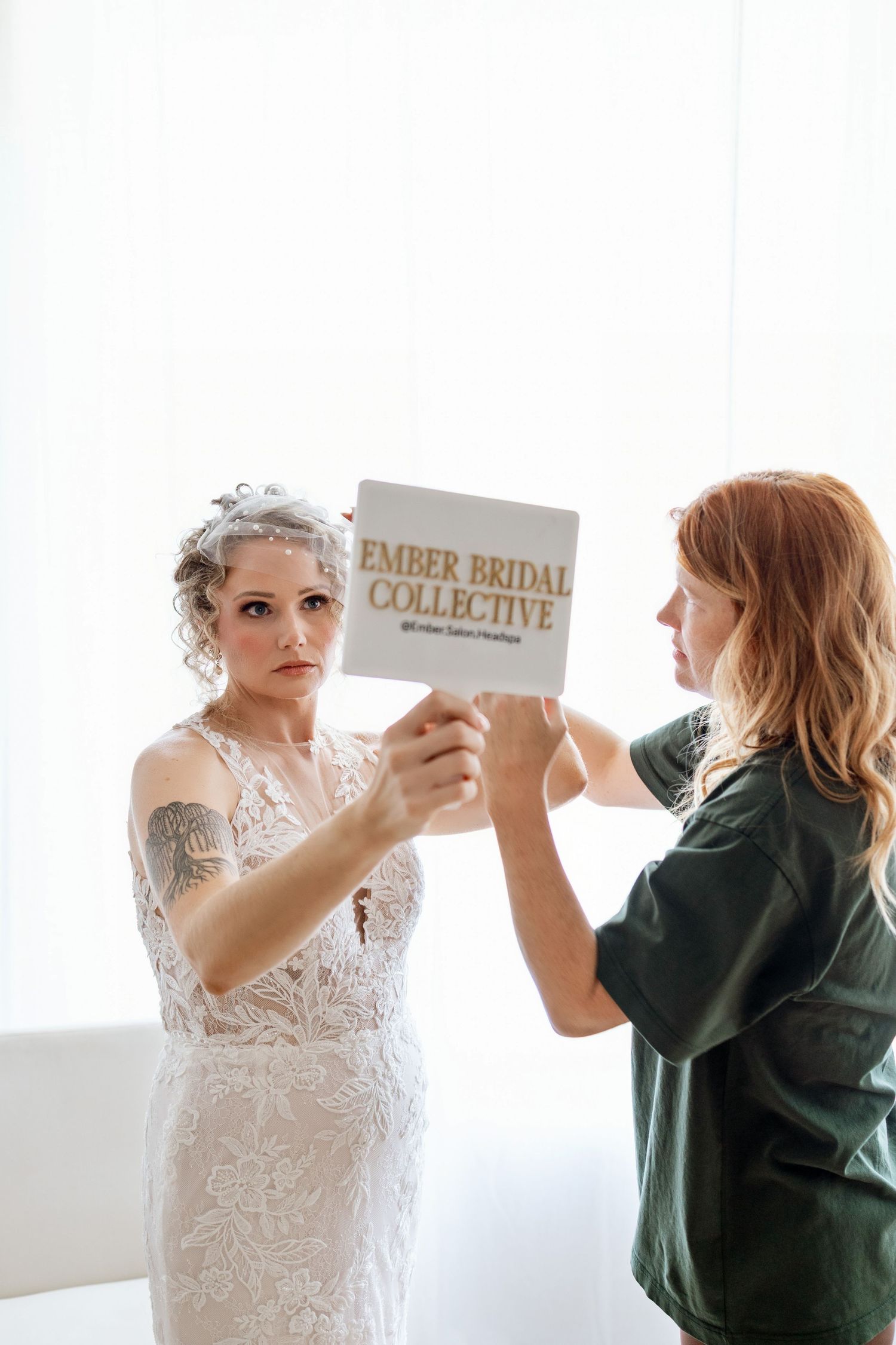 Bride in lace wedding dress holds Ember Bridal Collective sign with red-haired stylist in bright studio setting.