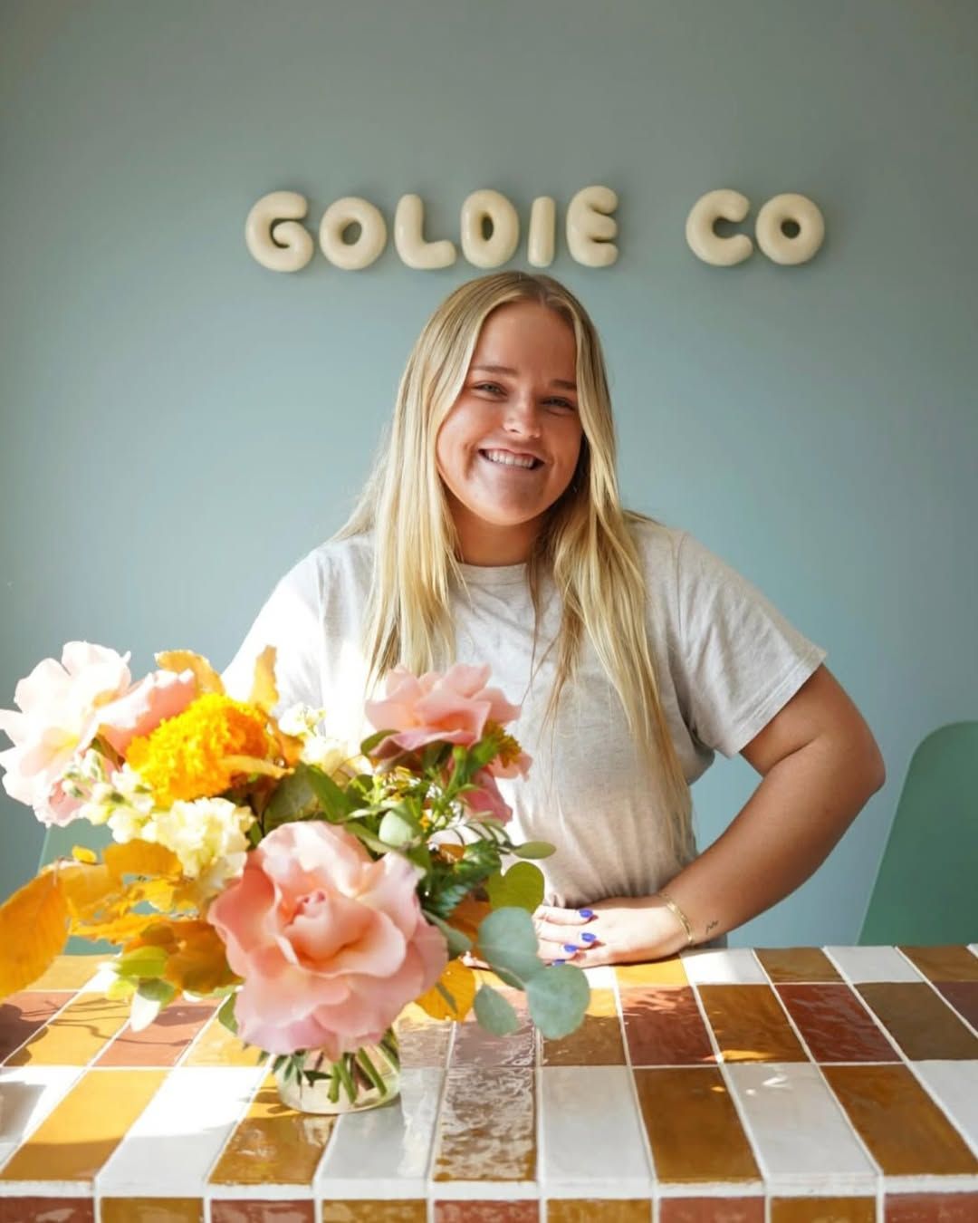 Smiling blonde woman standing behind a floral arrangement at Goldie Co, with pink and yellow flowers on a tiled table.