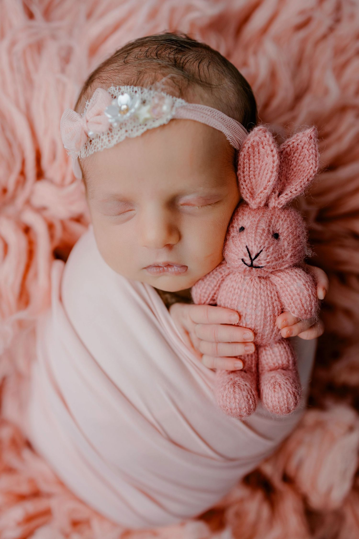 Sleeping newborn wrapped in white fabric holding a pink knitted bunny toy on coral-colored textured background.