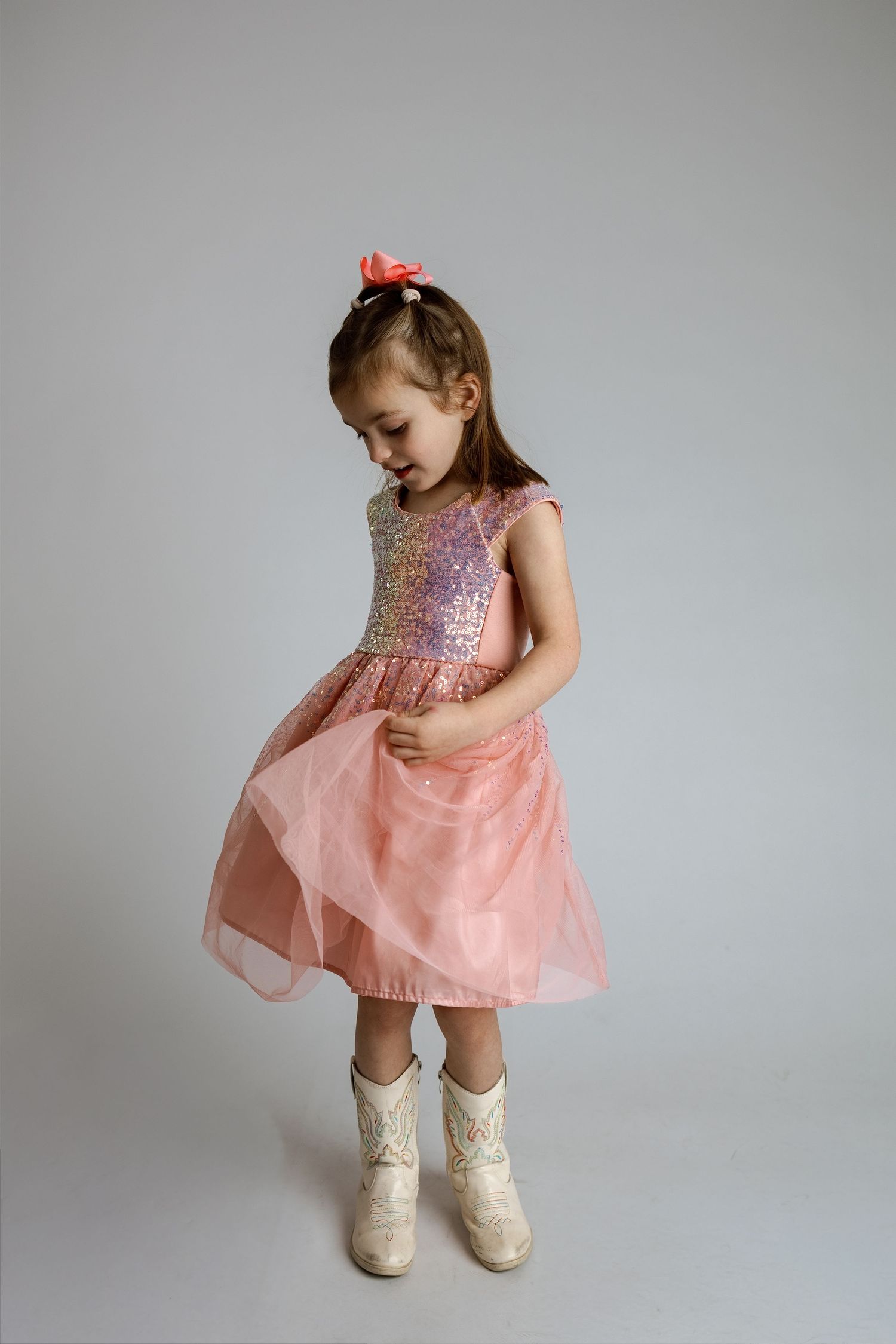 Young dancer in pink sequined dress and white boots poses gracefully against gray background.