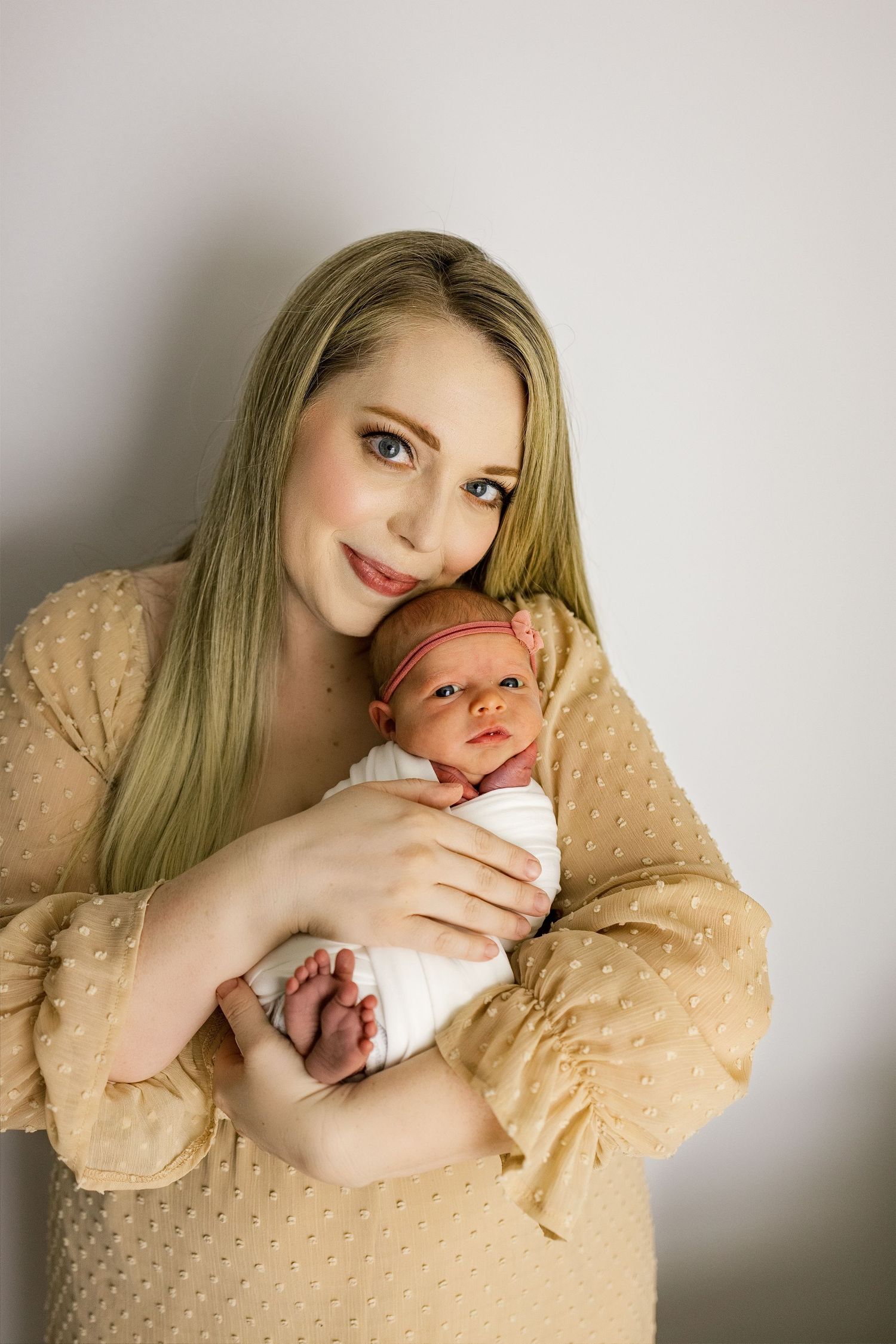 Mother in beige sweater holding newborn baby against white wall.