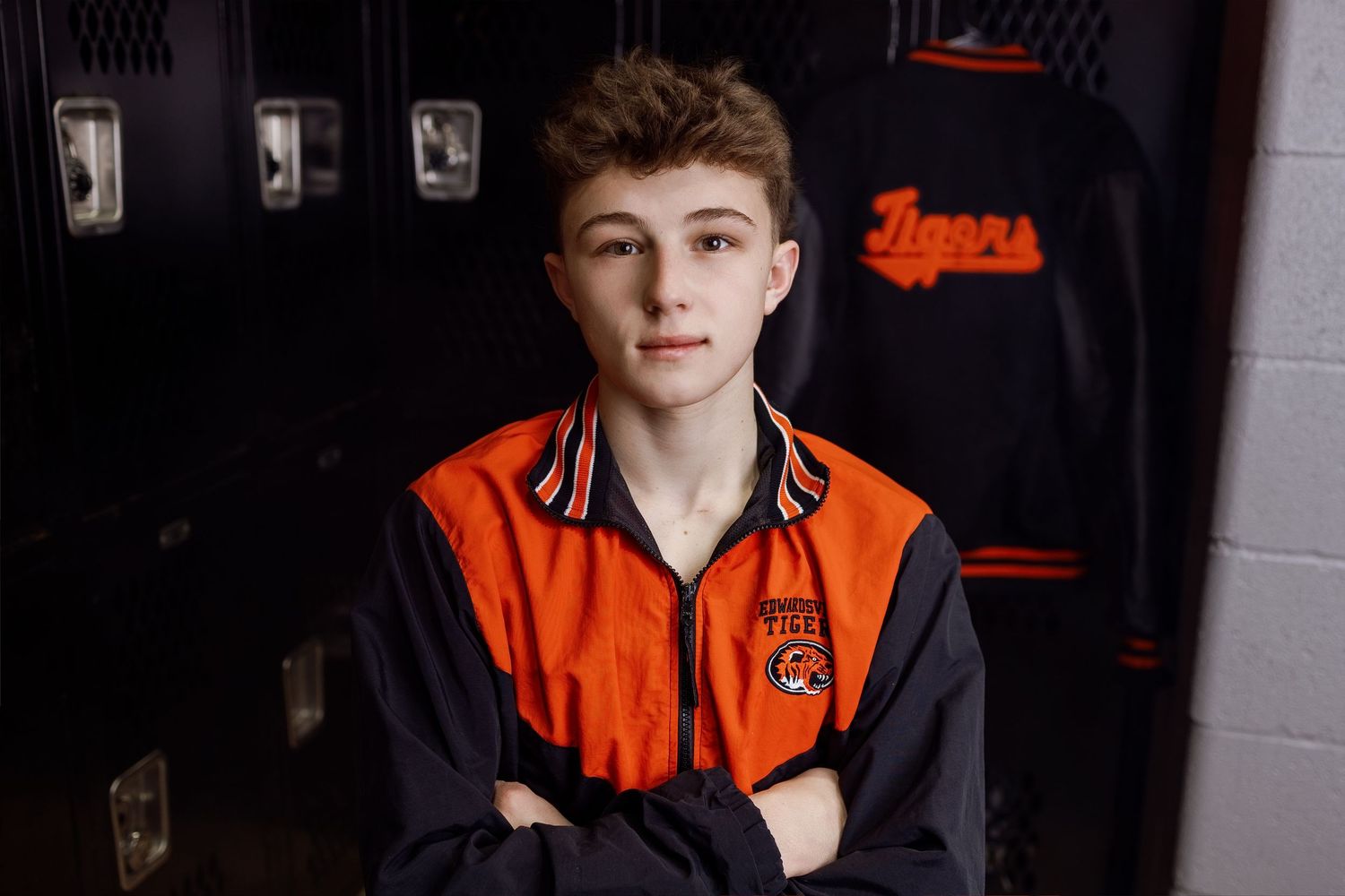 Young athlete in orange and black varsity jacket poses against dark background.