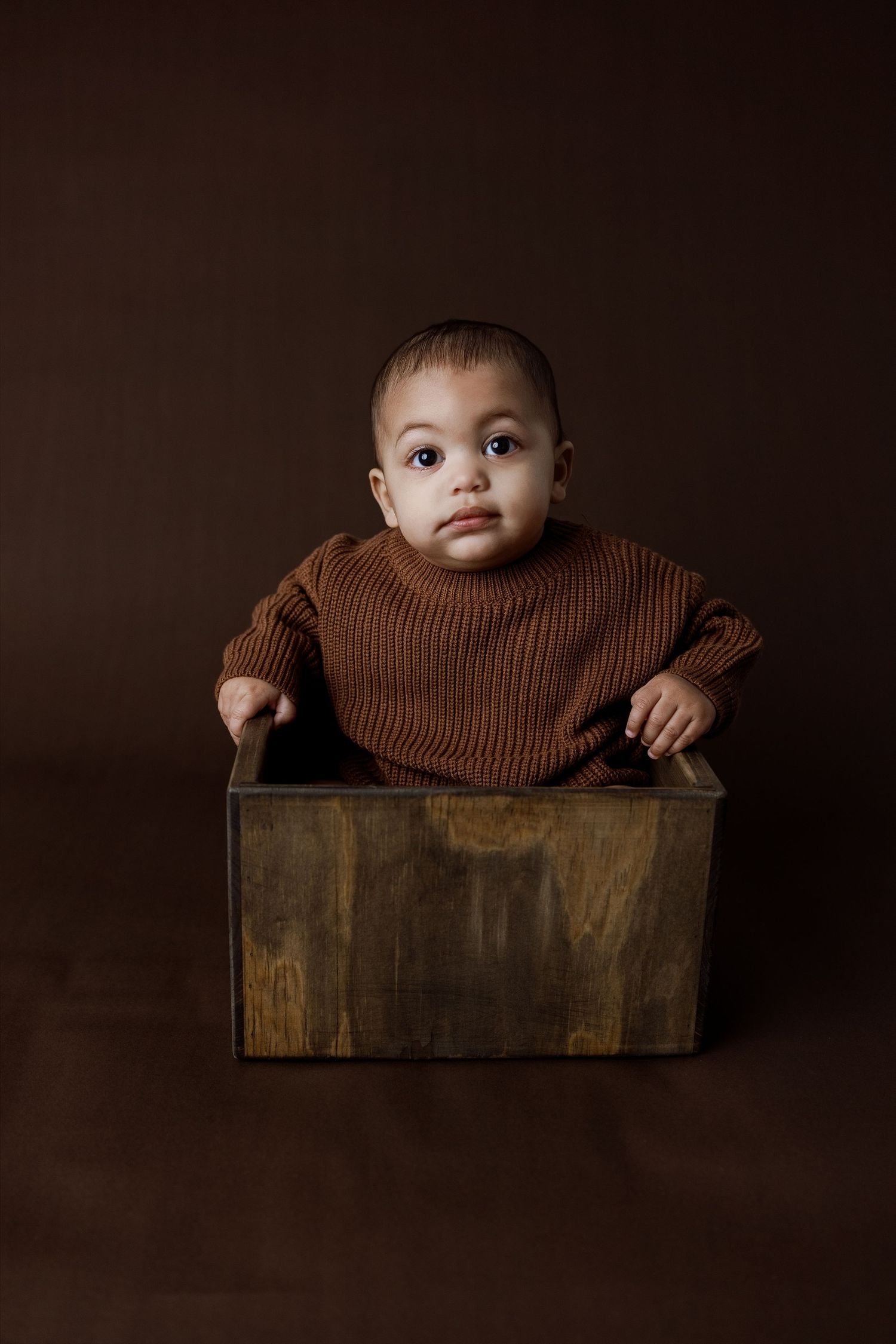 A small baby sits inside a rustic wooden box against a dark background in a series of artistic portraits.