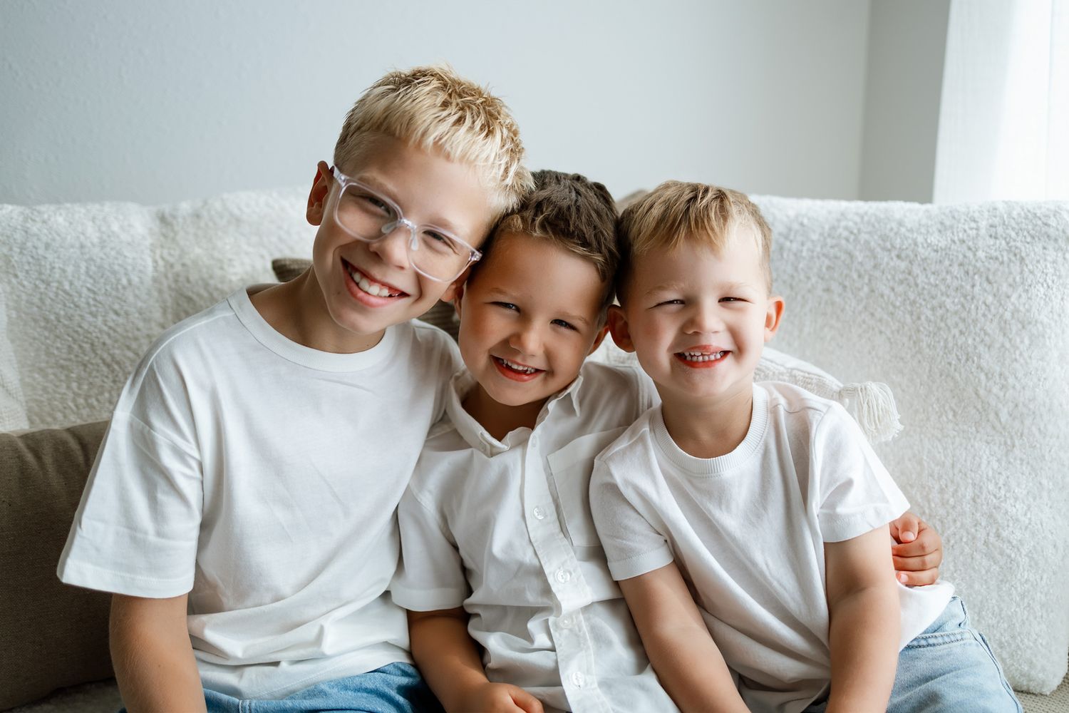 Three smiling boys in white t-shirts sitting together on a couch, sharing a warm embrace.