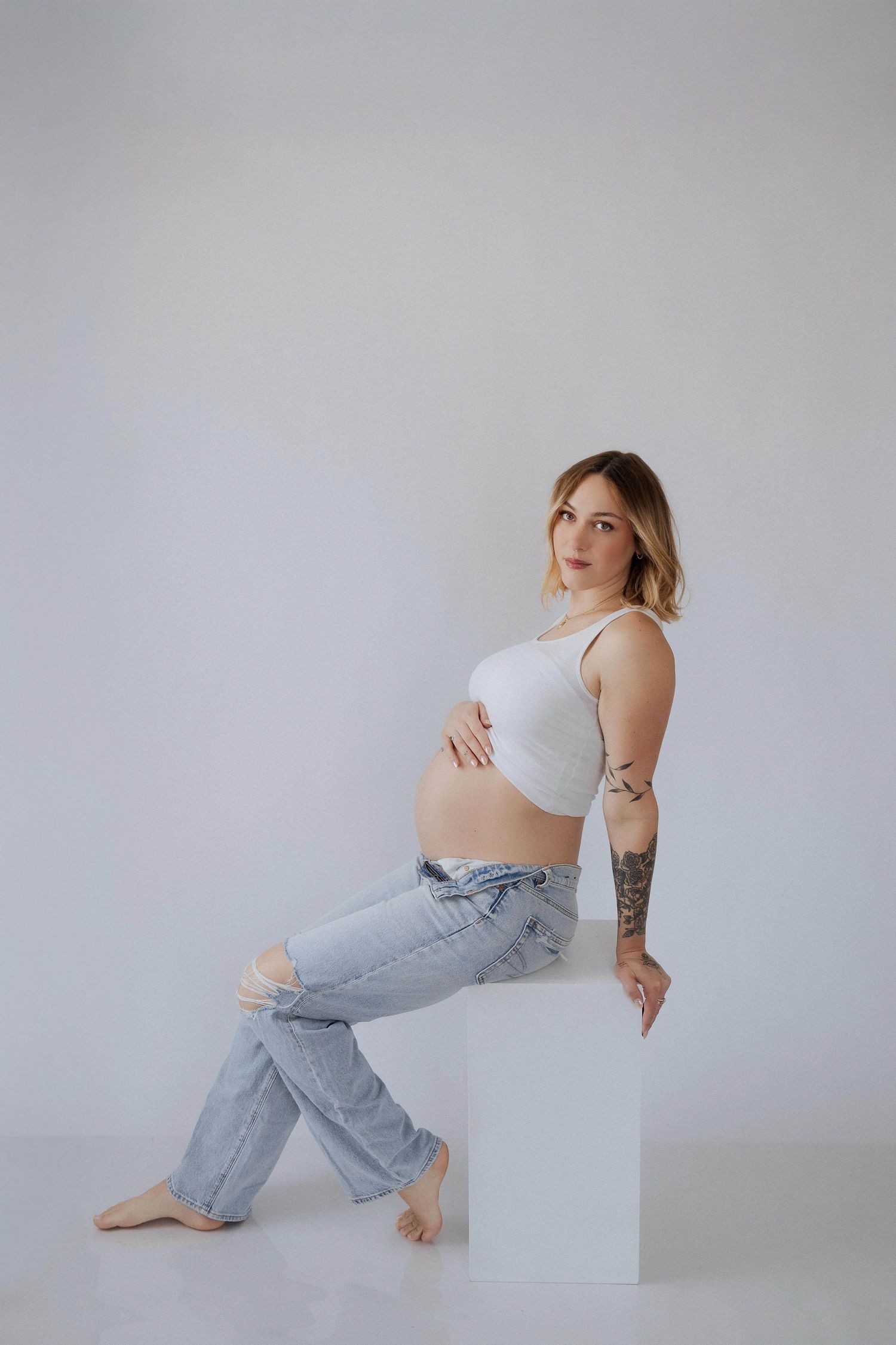 Pregnant woman in white crop top and distressed jeans sits barefoot on white cube against neutral background.
