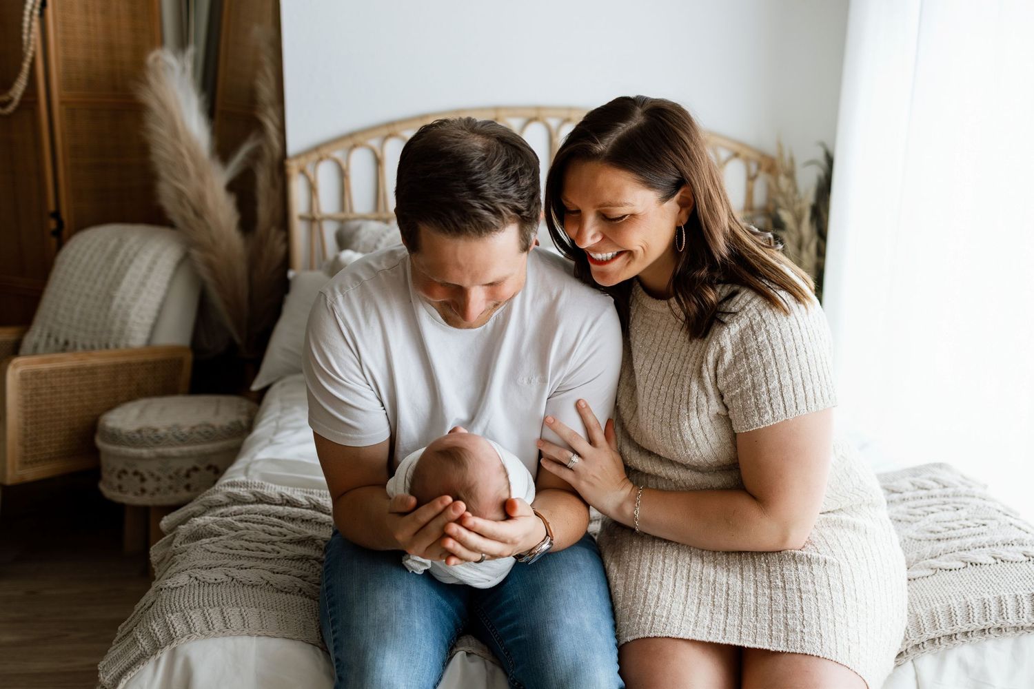 Happy parents sitting on a bed, lovingly cradling their newborn baby in a cozy, bohemian-styled nursery room.