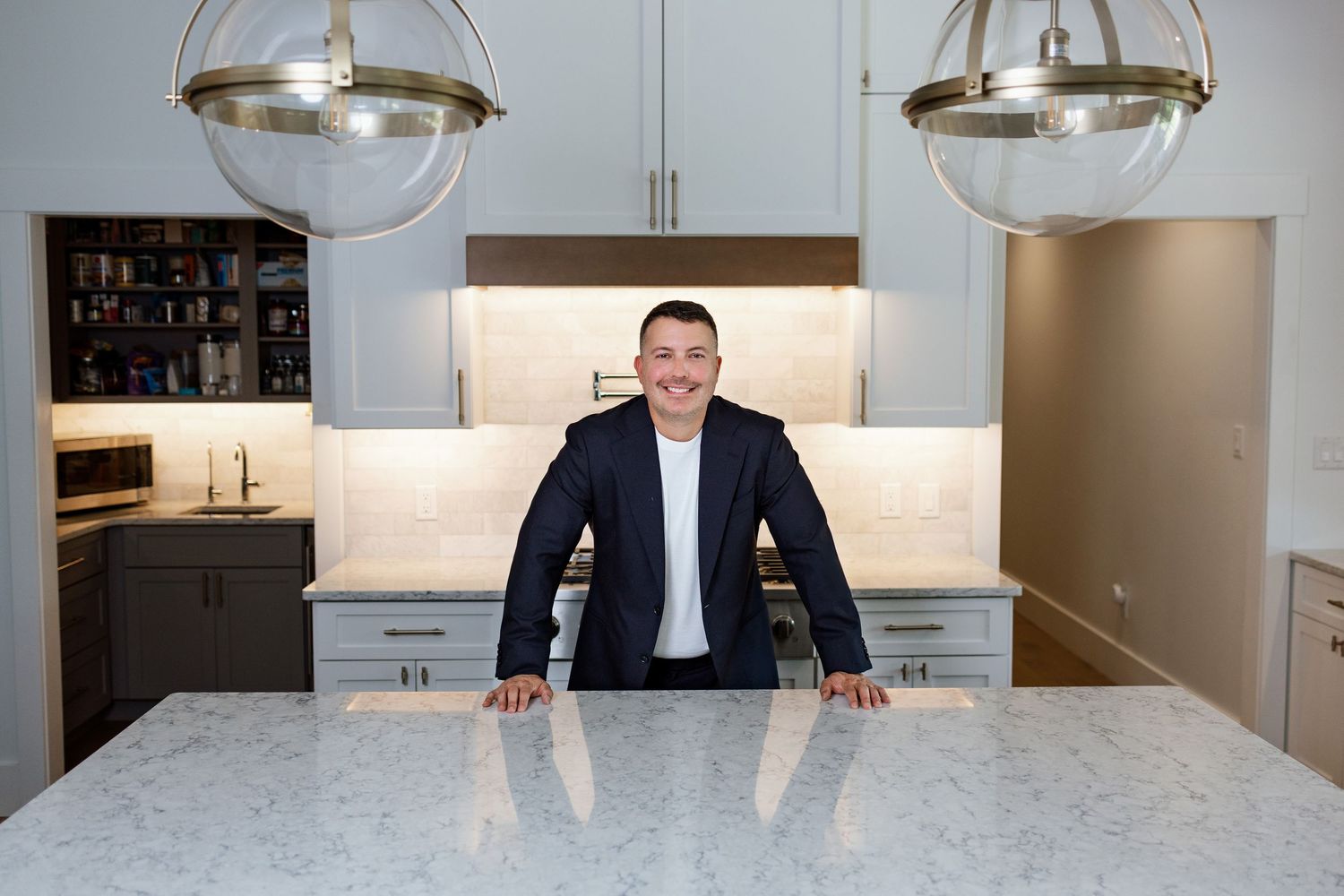 Man in blazer stands confidently at a marble kitchen island with pendant lights and gray cabinets.