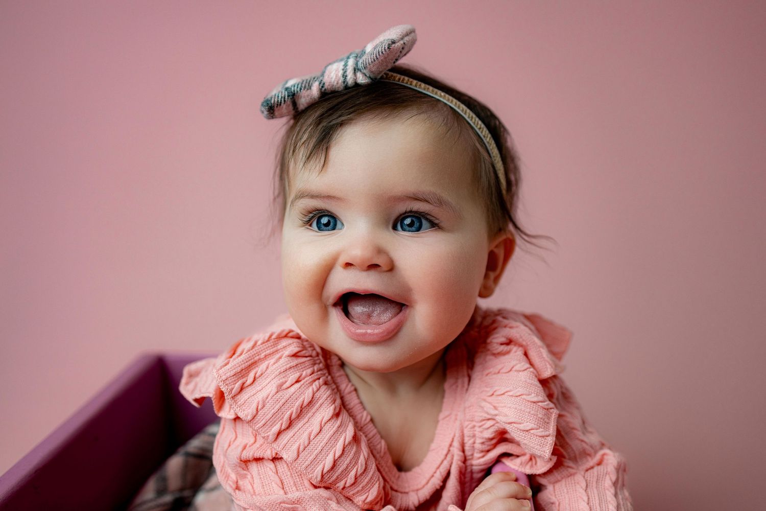 Joyful baby portrait on pink backdrop wearing peach colored ruffled dress and bow accessory.