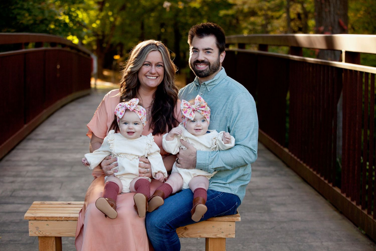 A couple sits with their twin babies on a wooden bench in a scenic outdoor setting.