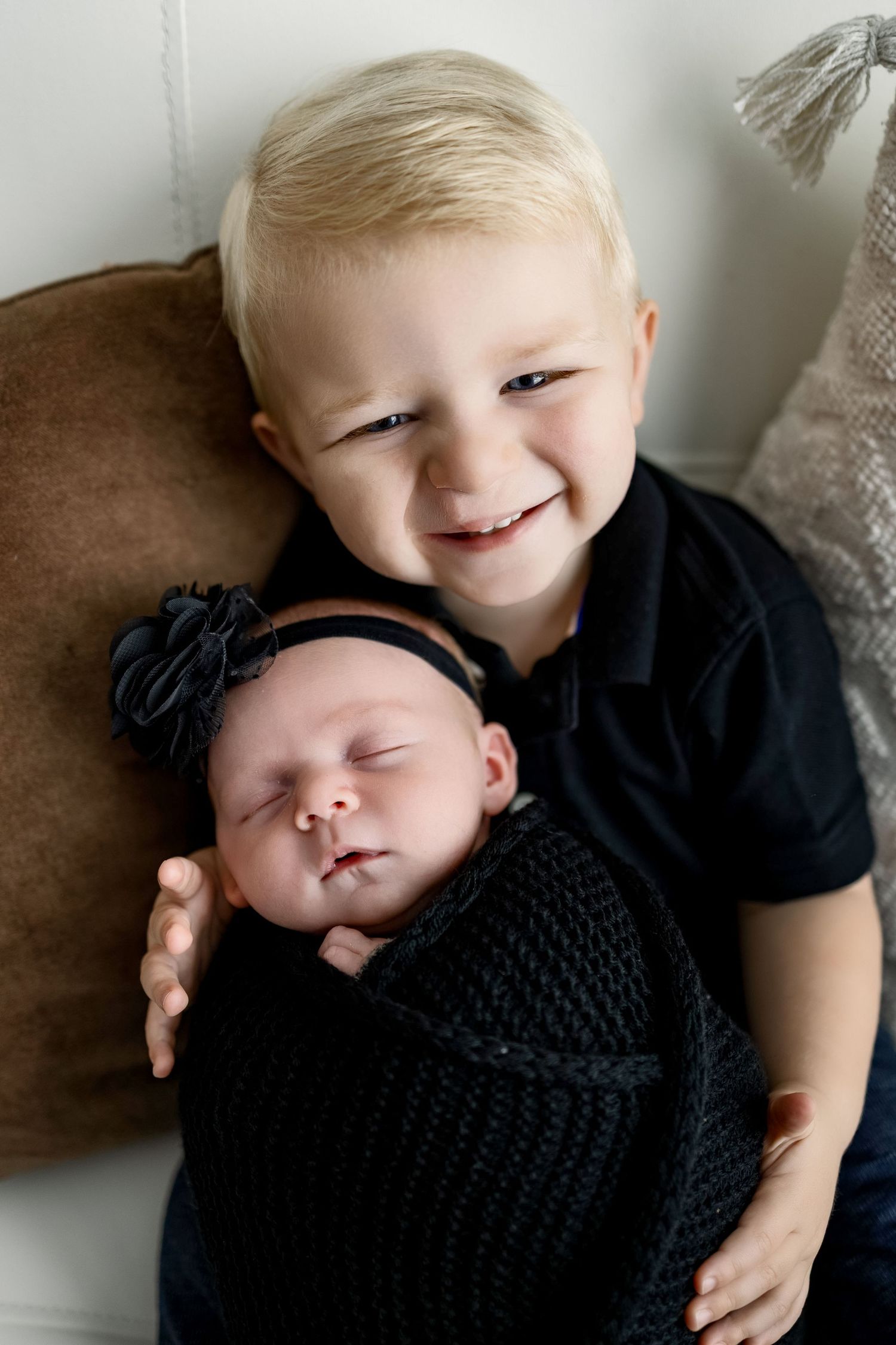 Two children in black outfits share a tender moment as the older one holds and smiles at the sleeping baby in knit wrap.