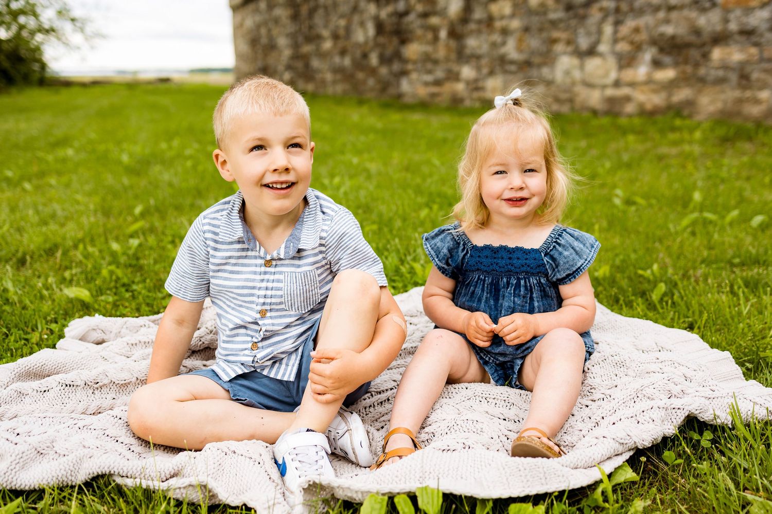 Two children sitting on a blanket on grass with an old stone wall in the background.