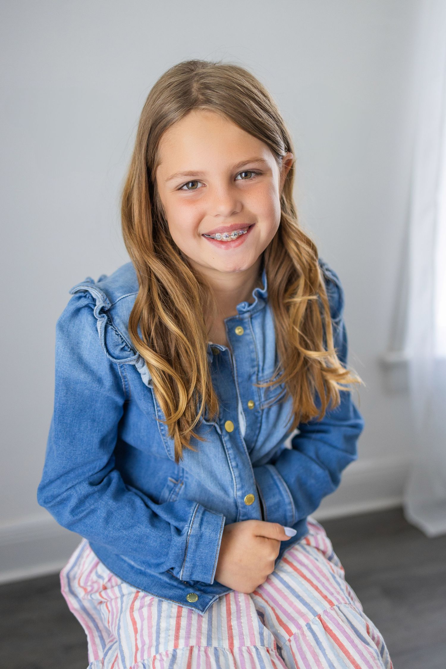 Young person wearing a blue denim jacket and striped pants sitting casually while smiling at the camera.
