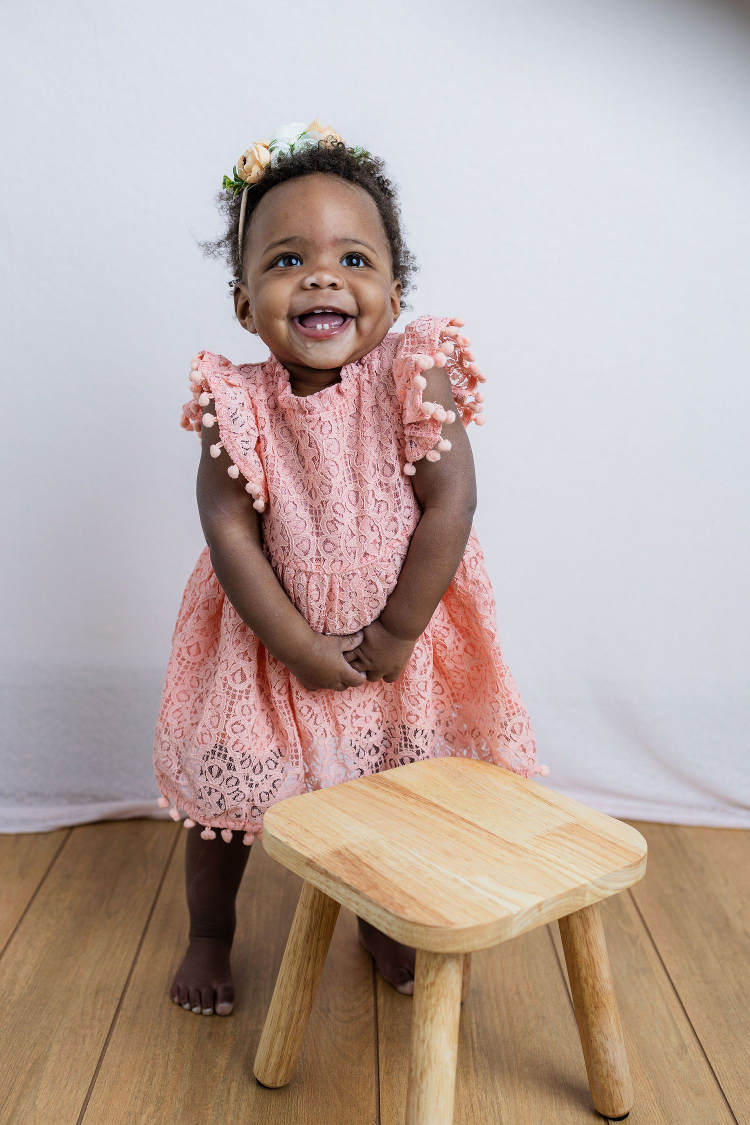 Joyful toddler in pink lace dress standing next to wooden stool against white backdrop.