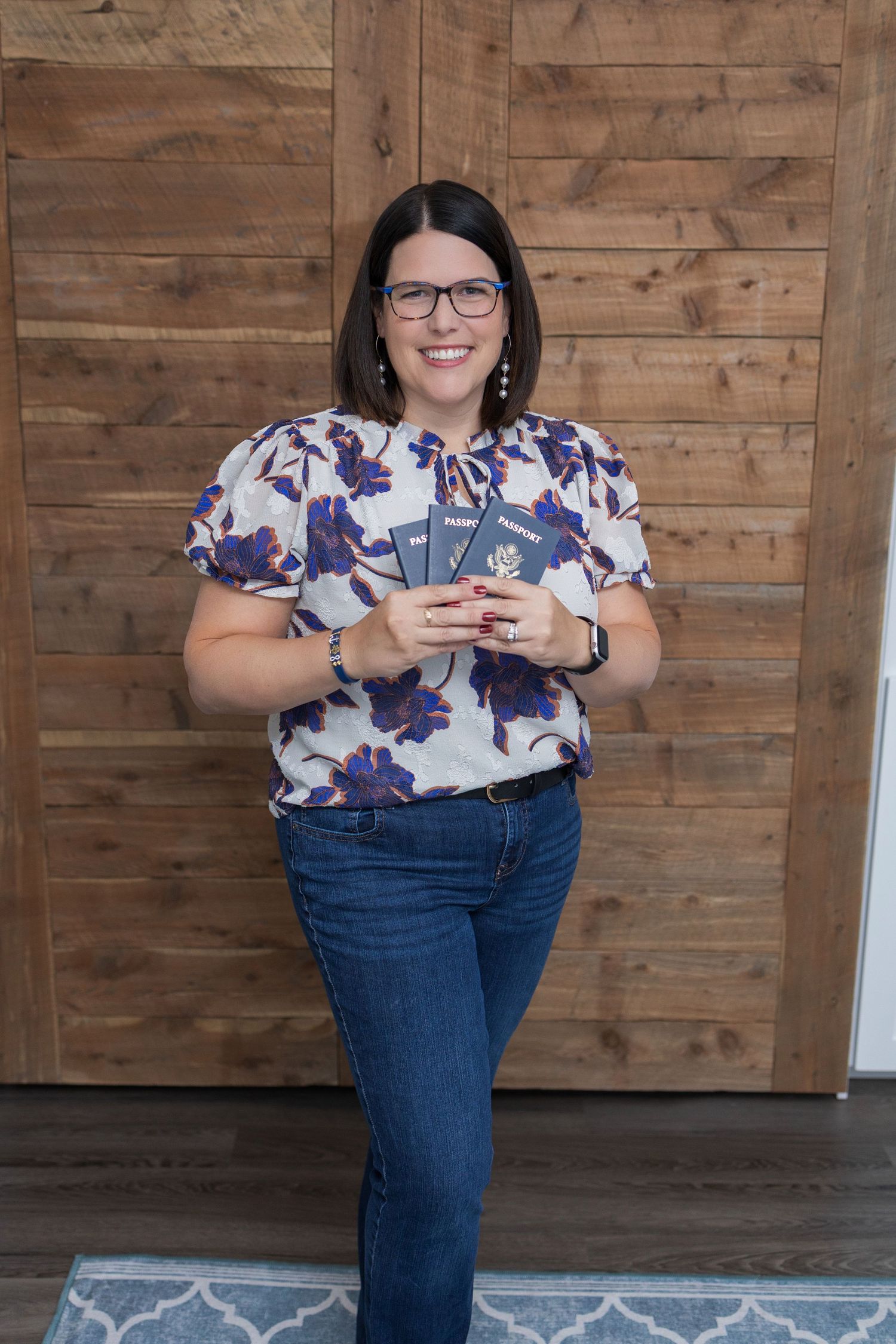 Person in floral top and jeans stands against wood panel wall while using smartphone in casual indoor setting.