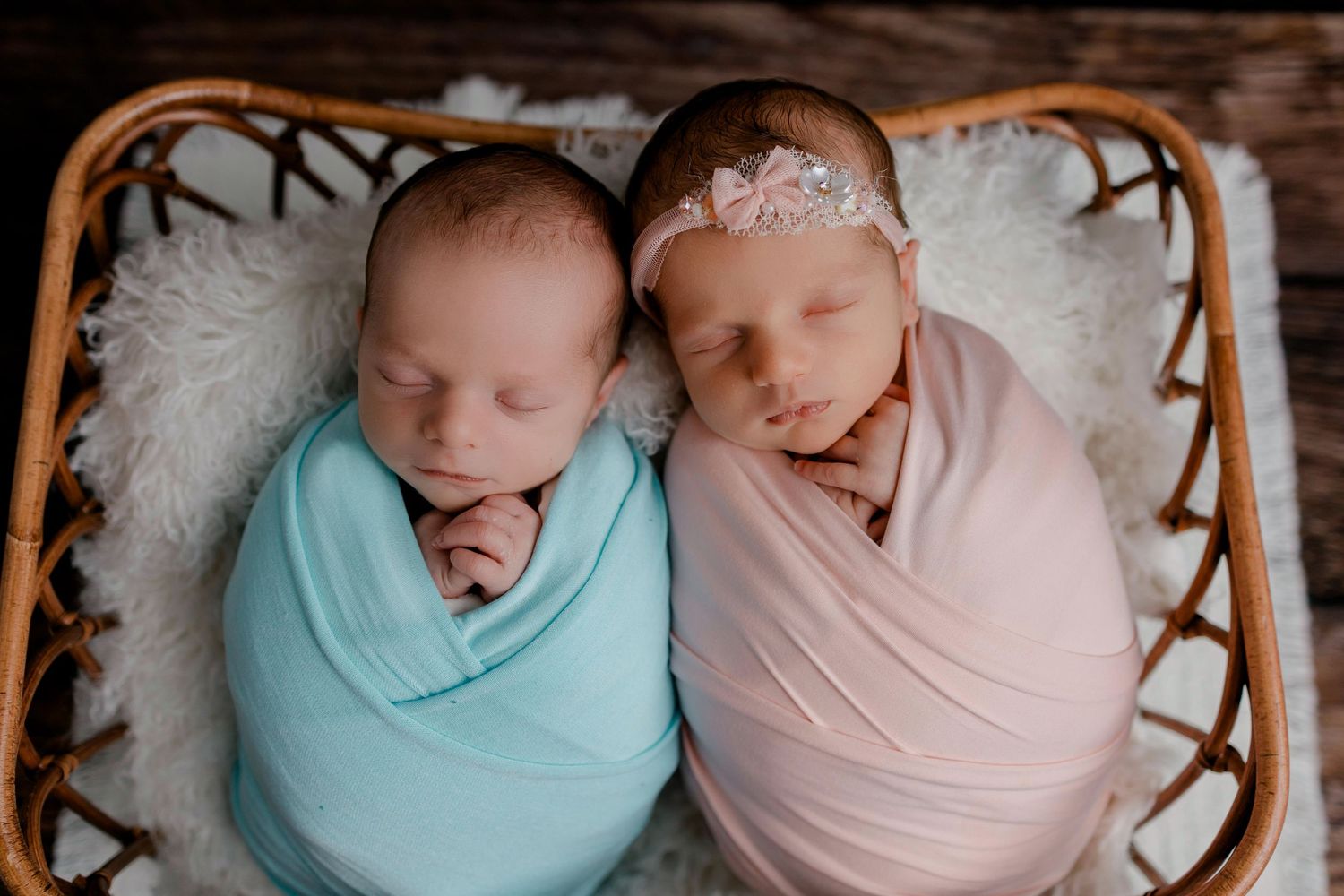 Sleeping newborn twins swaddled in soft blue and pink blankets nestled together in woven basket.