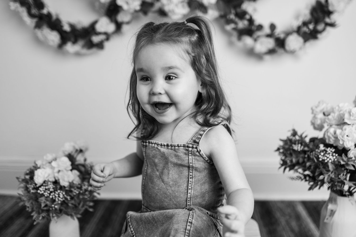 A playful and joyful moment is captured in black and white as a child laughs with pure delight near floral arrangements.