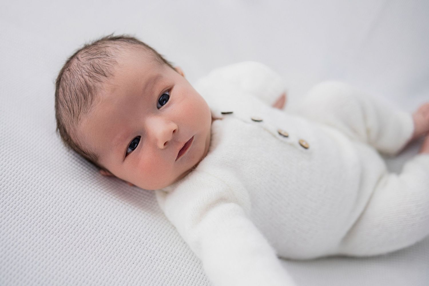 Adorable infant wearing a soft white cardigan lies on a textured gray blanket looking at the camera.