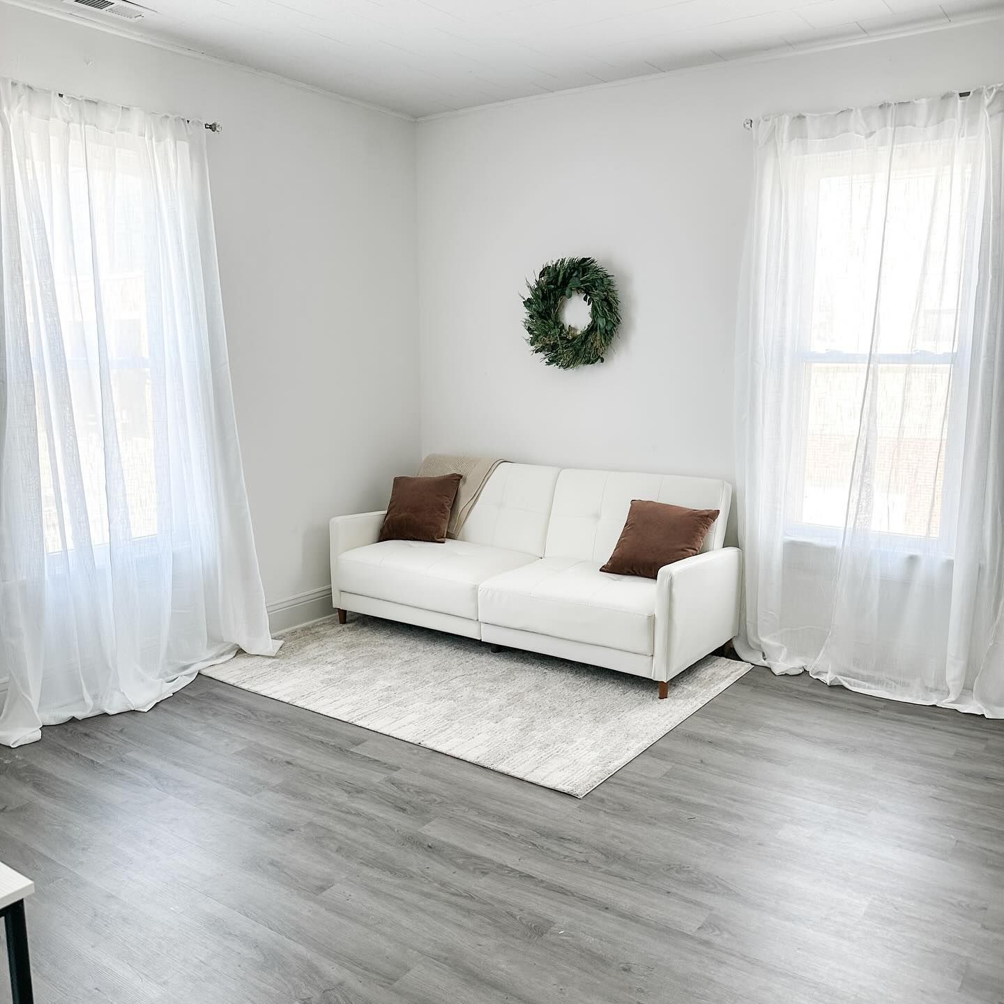 Clean white room with sofa, wreath and sheer curtains on grey flooring.