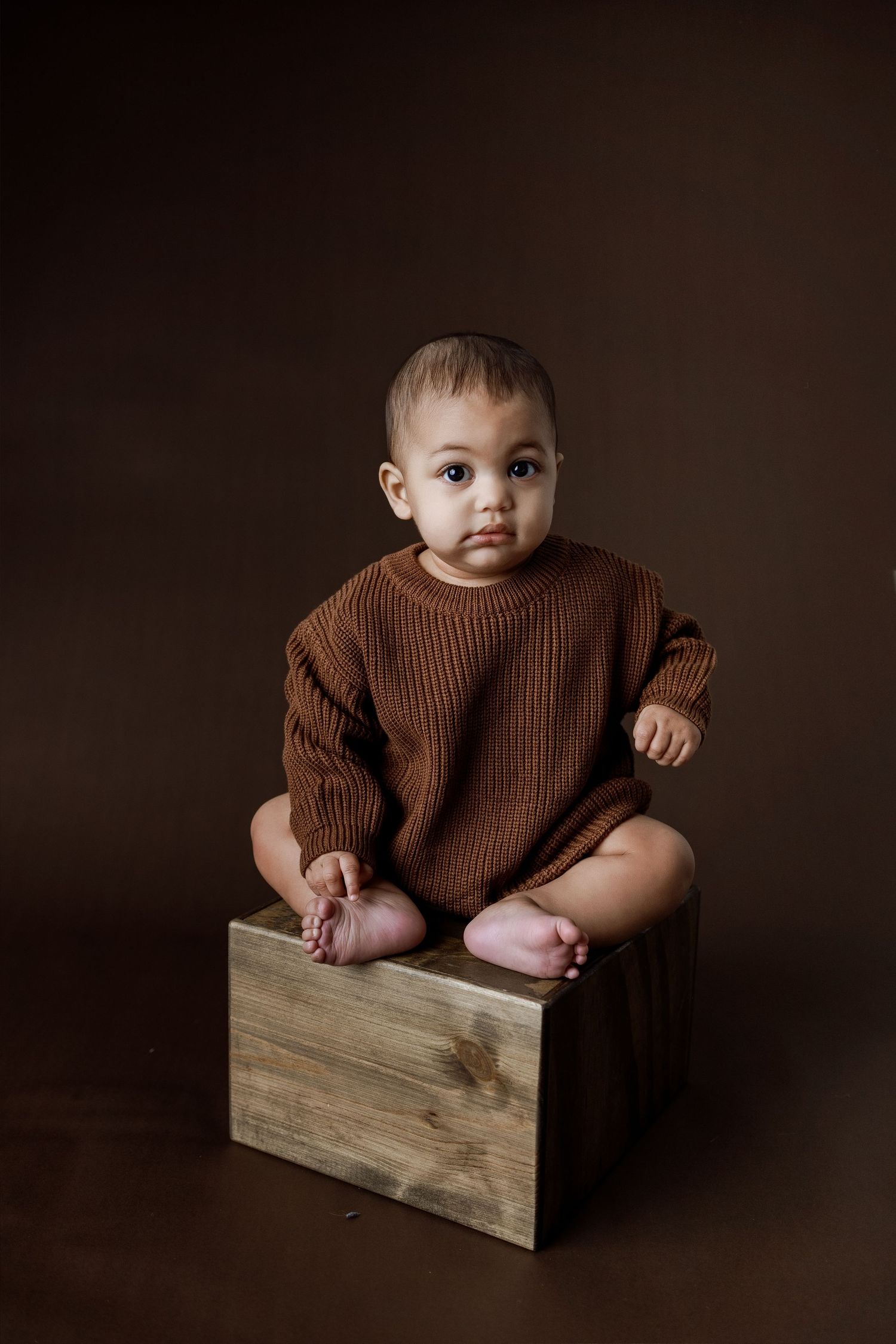 A baby in a brown sweater sits in a wooden box against a dark background in a series of artistic portraits.