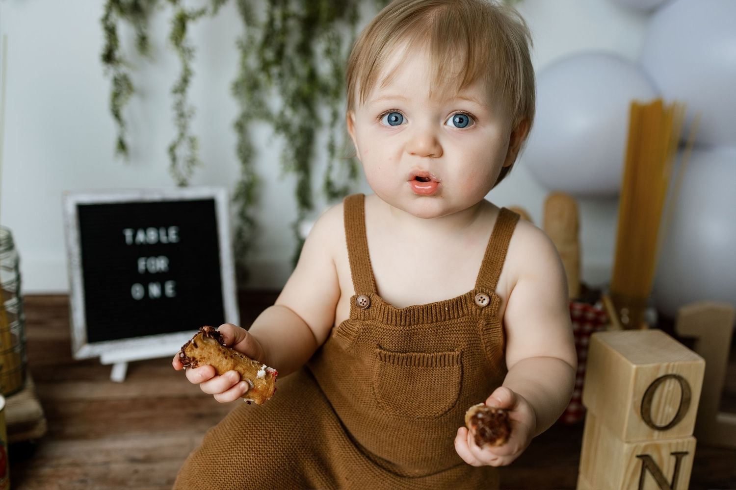A child in brown overalls sits at a table with chocolate cake and frosting smeared on their face.