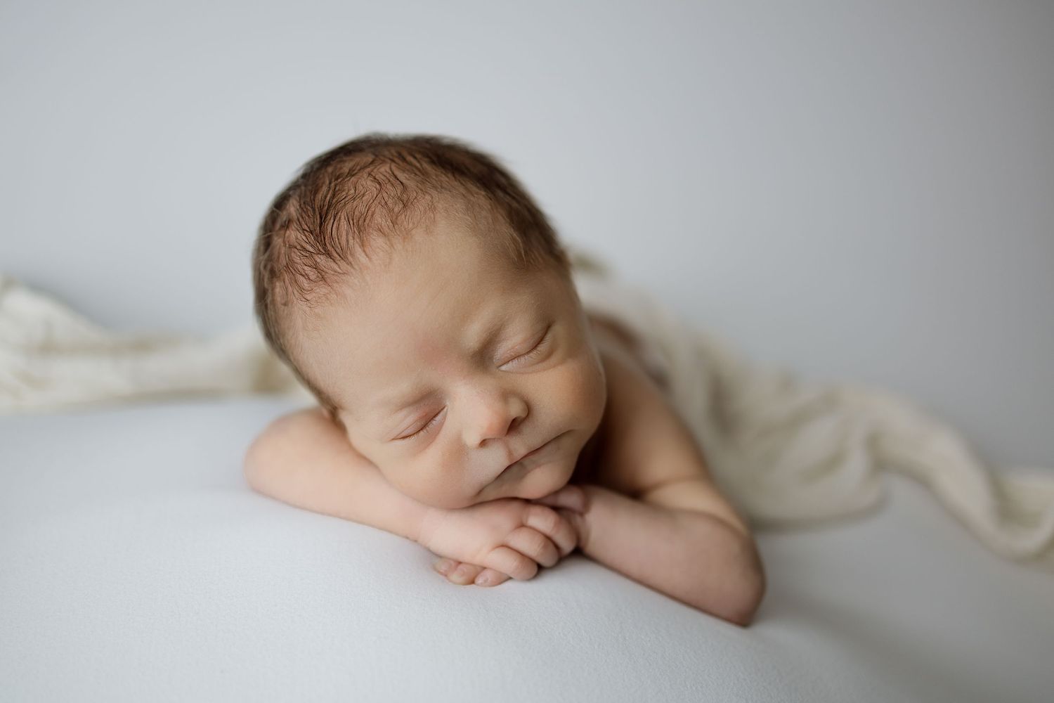 A sleeping newborn baby wrapped in white fabric rests peacefully against a light background.