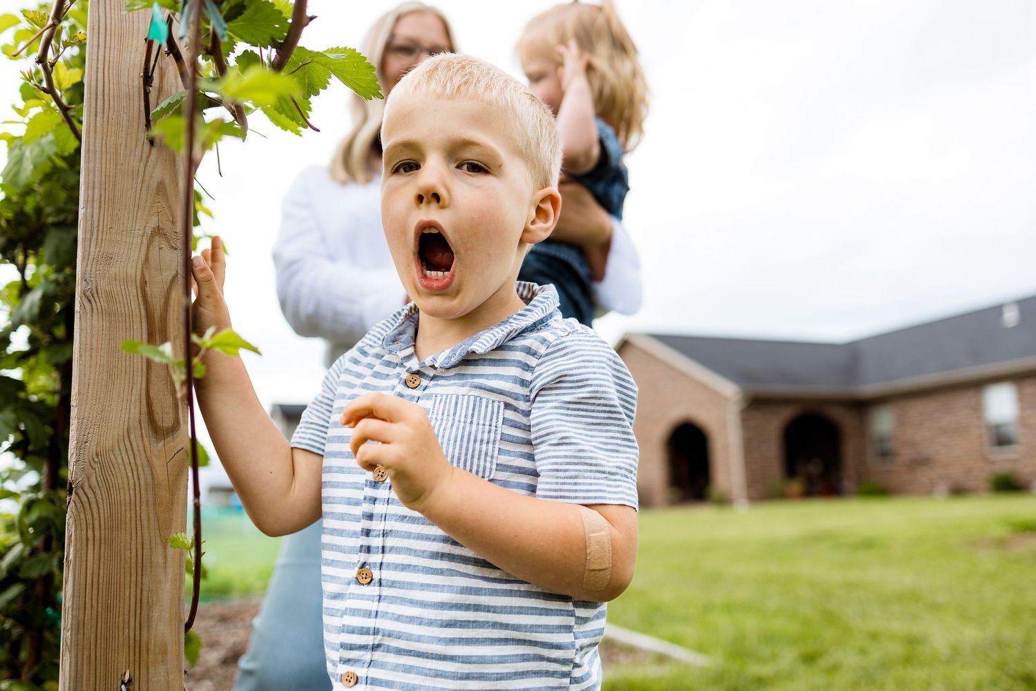 Young child in striped shirt yawns while playing outdoors in a green yard with a brick building in background.