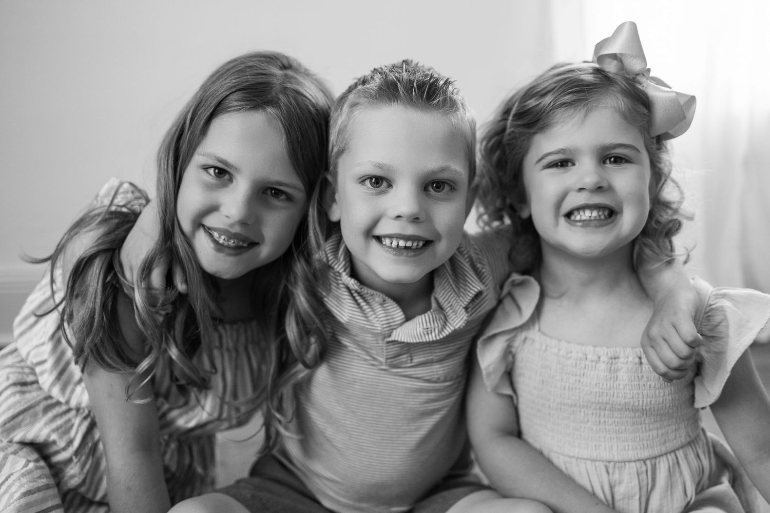 A black and white portrait of three young children sitting together and smiling joyfully at the camera.