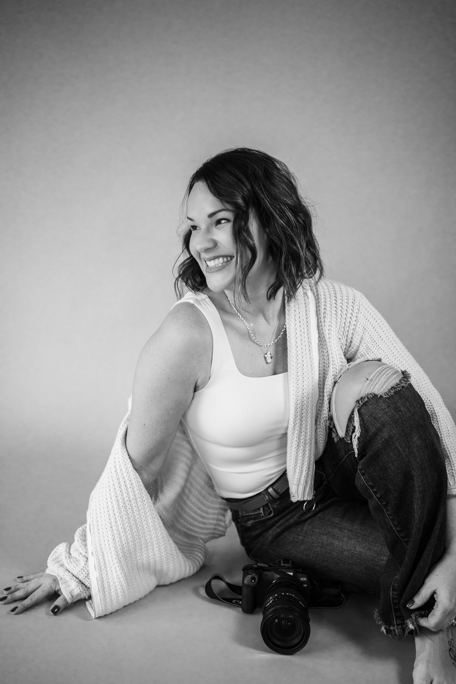 Black and white photo of a smiling woman sitting on floor holding a camera, wearing jeans and a cardigan.