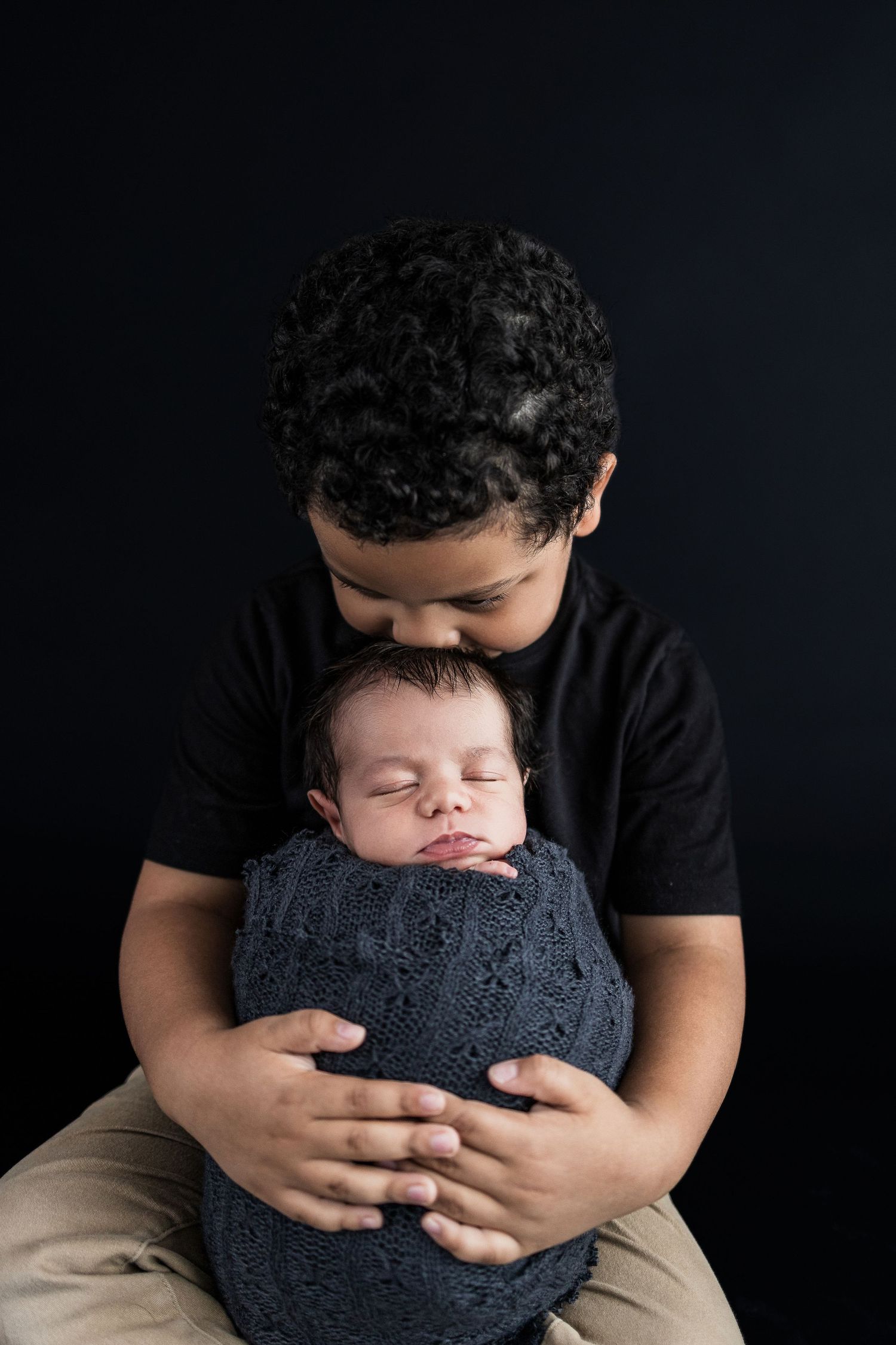 A young child holds a sleeping newborn wrapped in a gray knit blanket against a dark background.
