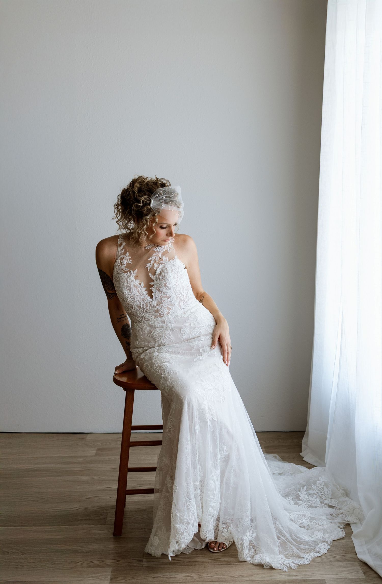 Bride in lace mermaid wedding gown with floral headpiece seated on wooden stool near sheer curtains.