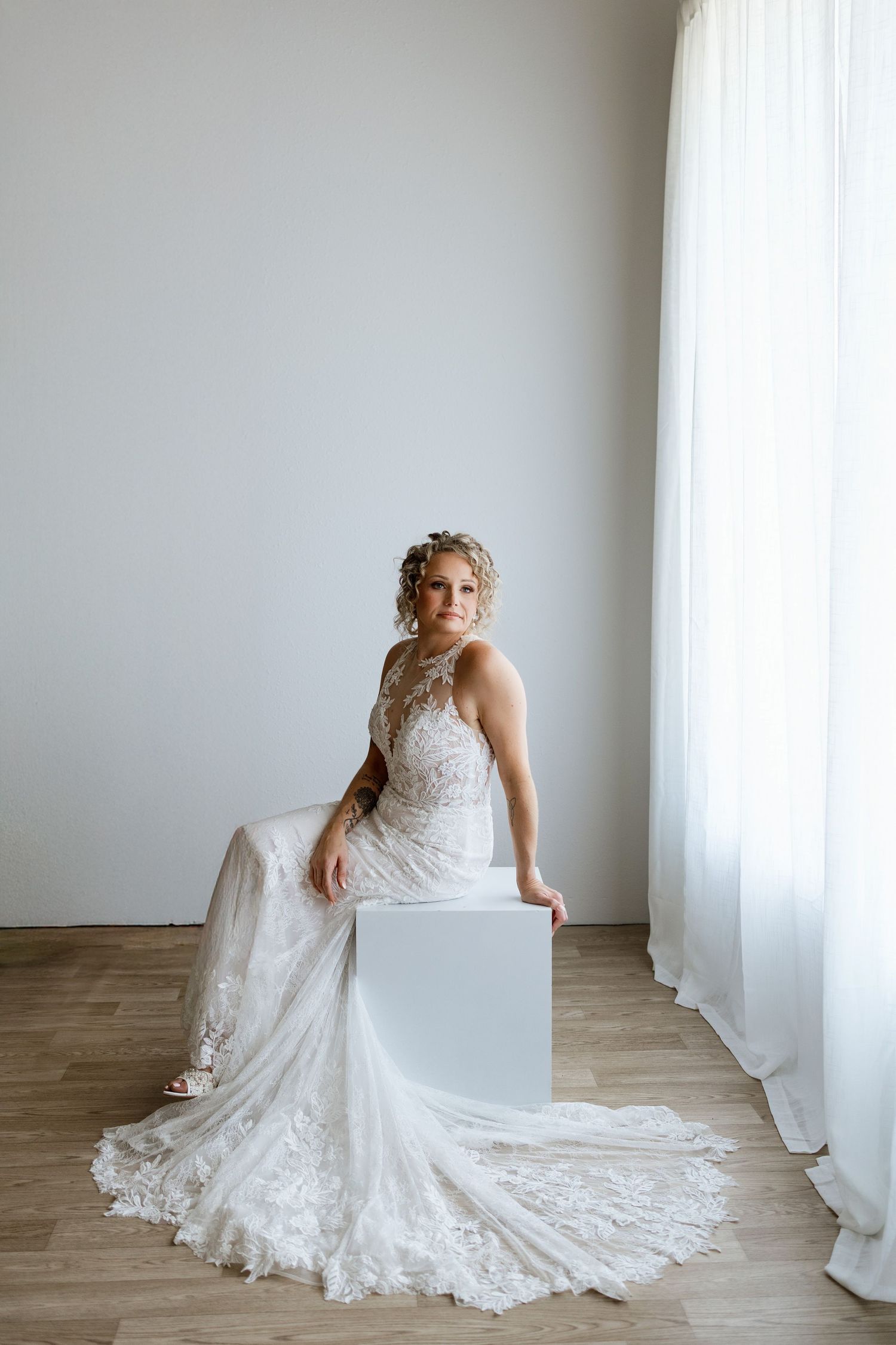 Bride in elegant lace wedding gown seated on white cube near sheer curtains in bright minimalist room.