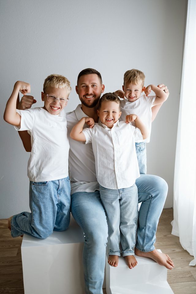 Father and three young sons in white shirts and jeans flexing muscles together in a bright white room.