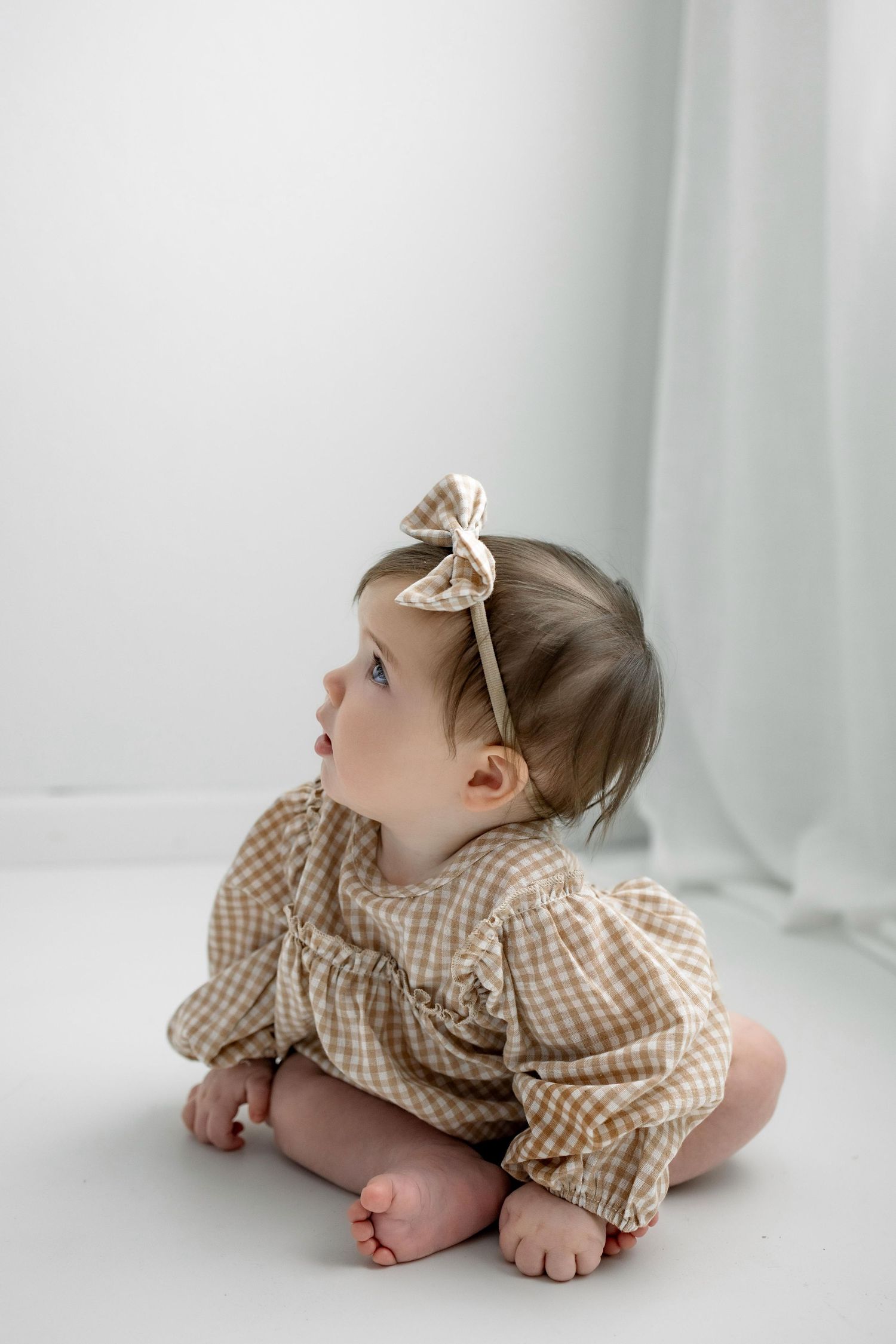 Baby in gingham dress and bow sits on white backdrop looking upward during studio photoshoot.