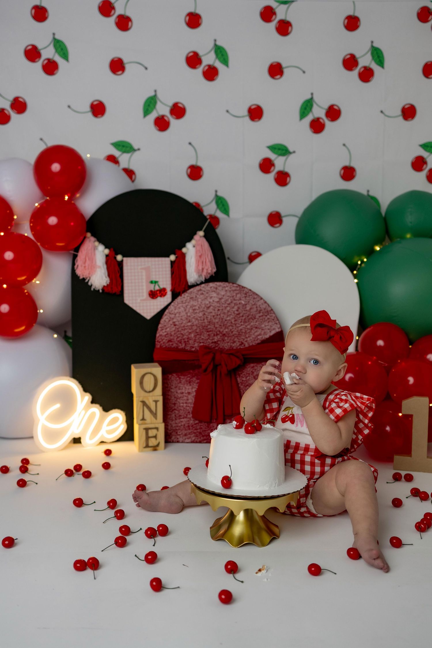 Cherry-themed first birthday cake smash photo session with red and green balloon decorations against white backdrop.