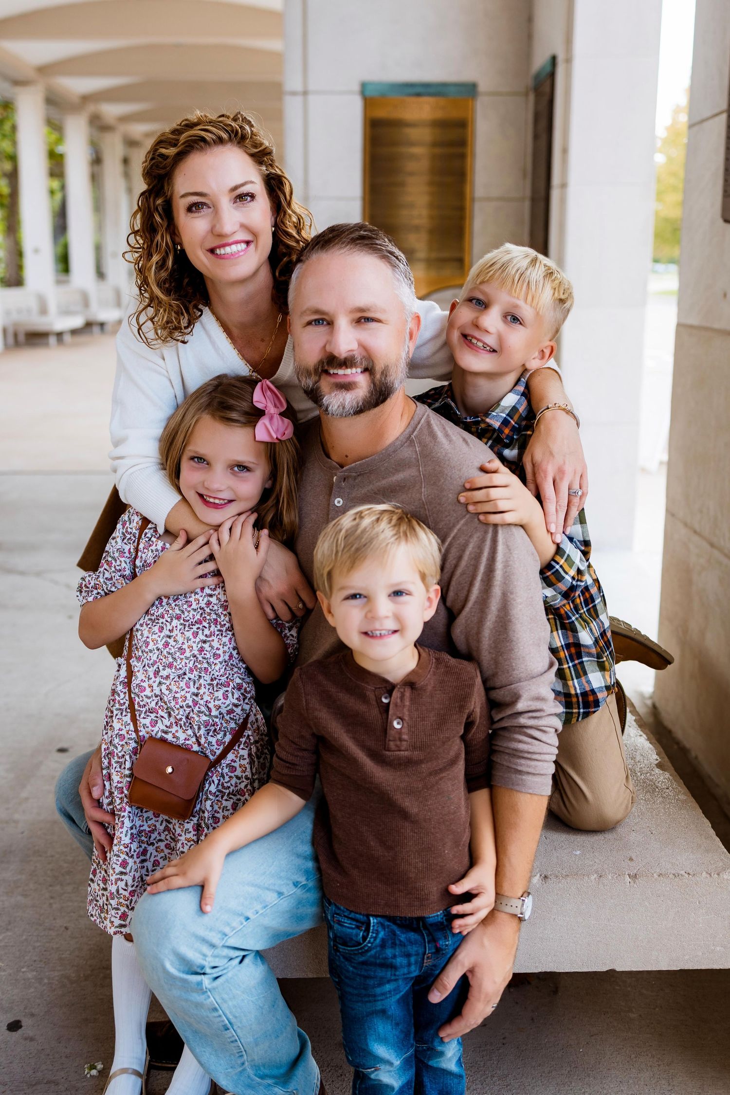 A family poses together in a covered outdoor walkway wearing casual clothing in matching brown and blue tones.