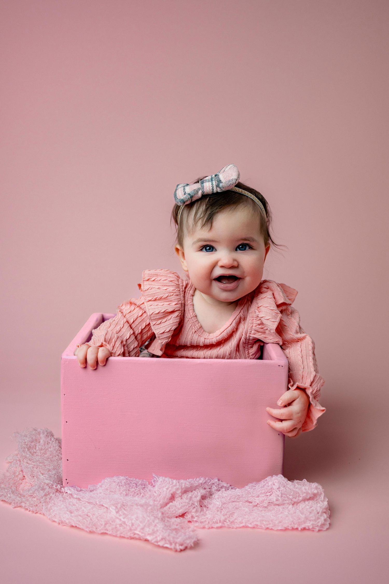 Joyful baby in pink ruffled dress poses inside pink box against rosy background.