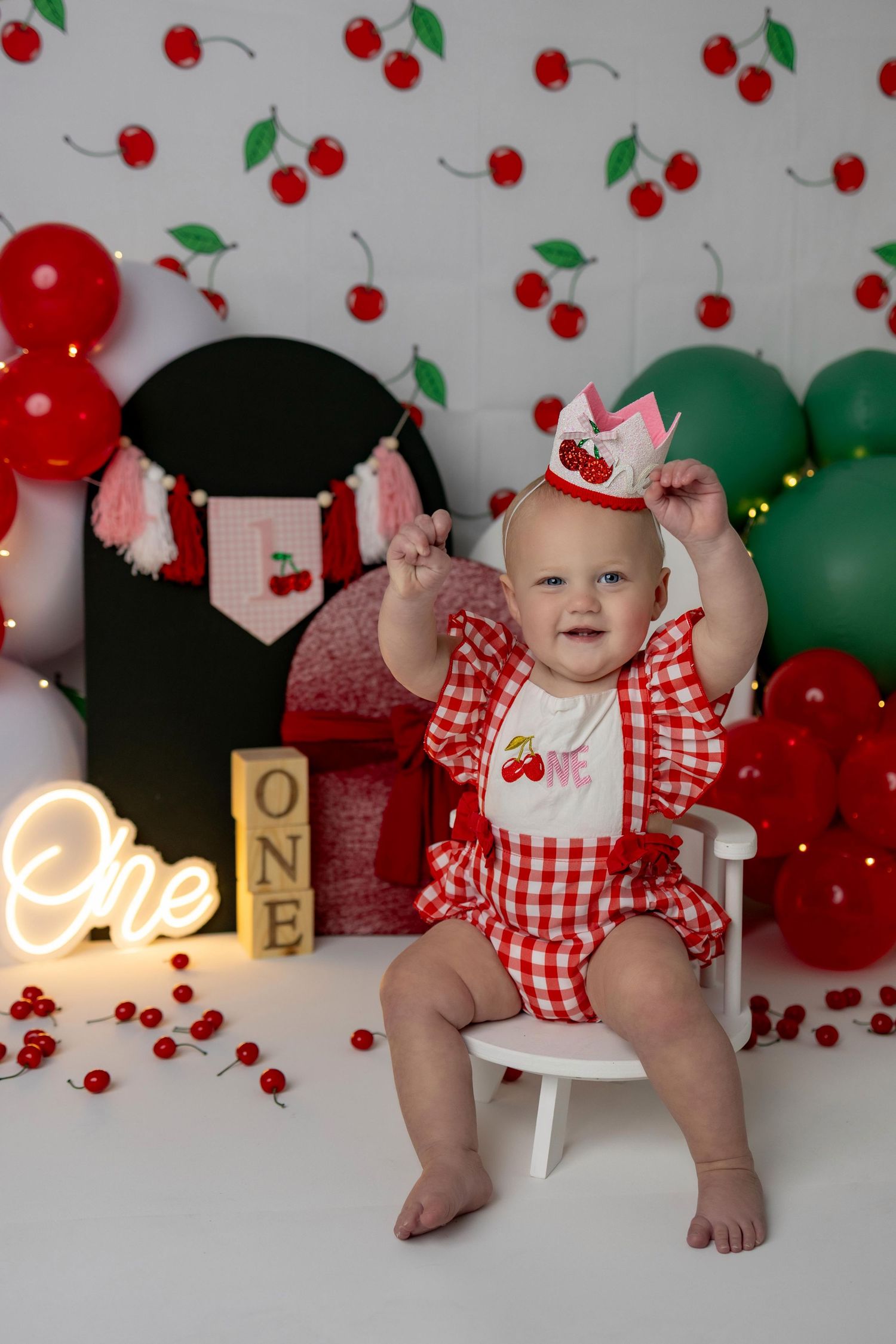 Baby in red gingham outfit sits among cherry-themed party decorations with balloons and festive decor.