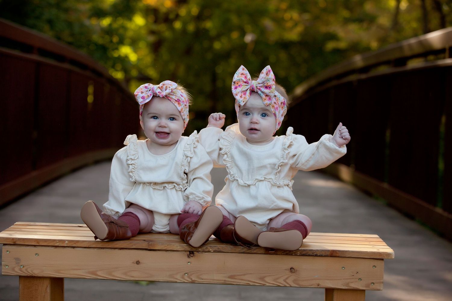 Two babies in matching white dresses and floral headbands sit together on a wooden bench in an outdoor setting.