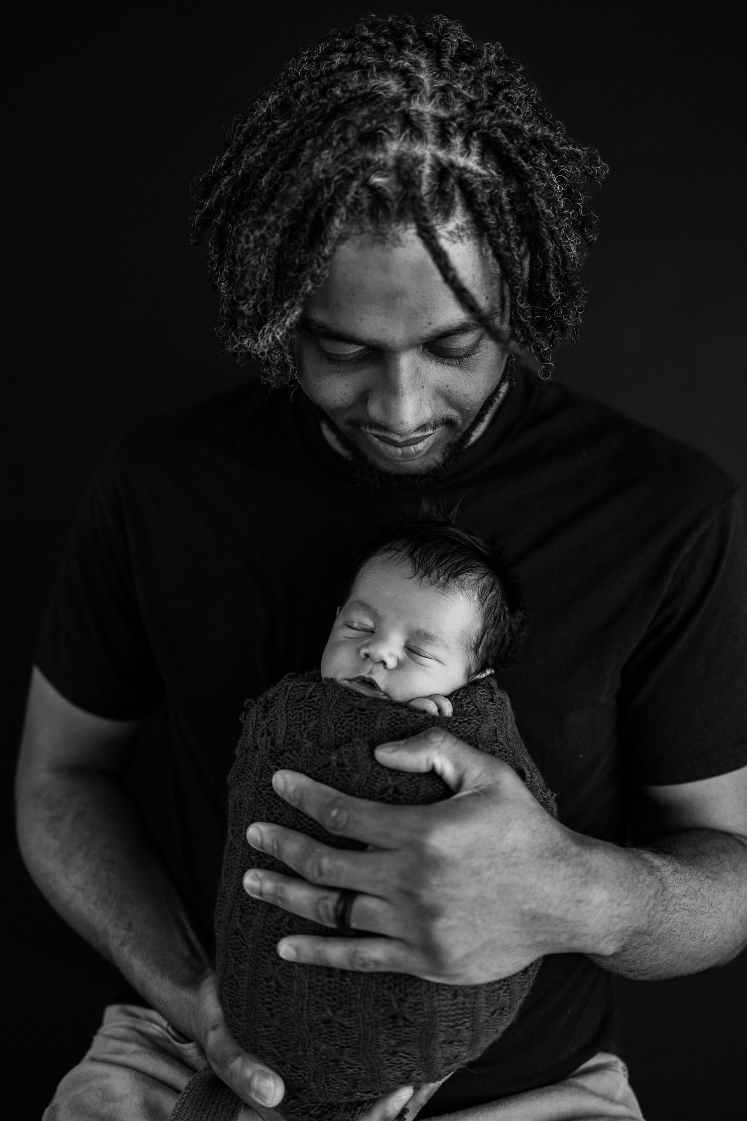 A tender black and white portrait of someone cradling a sleeping newborn swaddled in textured fabric.