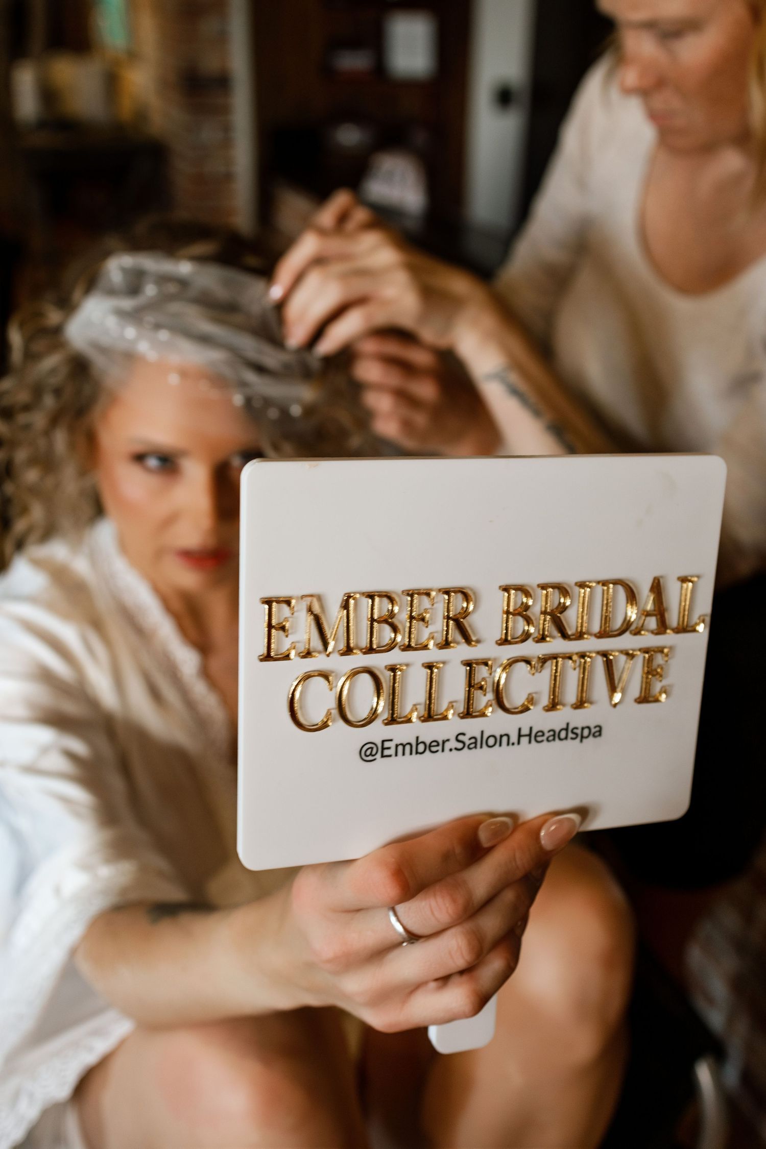 Bride holds Ember Bridal Collective sign while getting hair styled at @Ember.Salon.Headspa bridal beauty session.