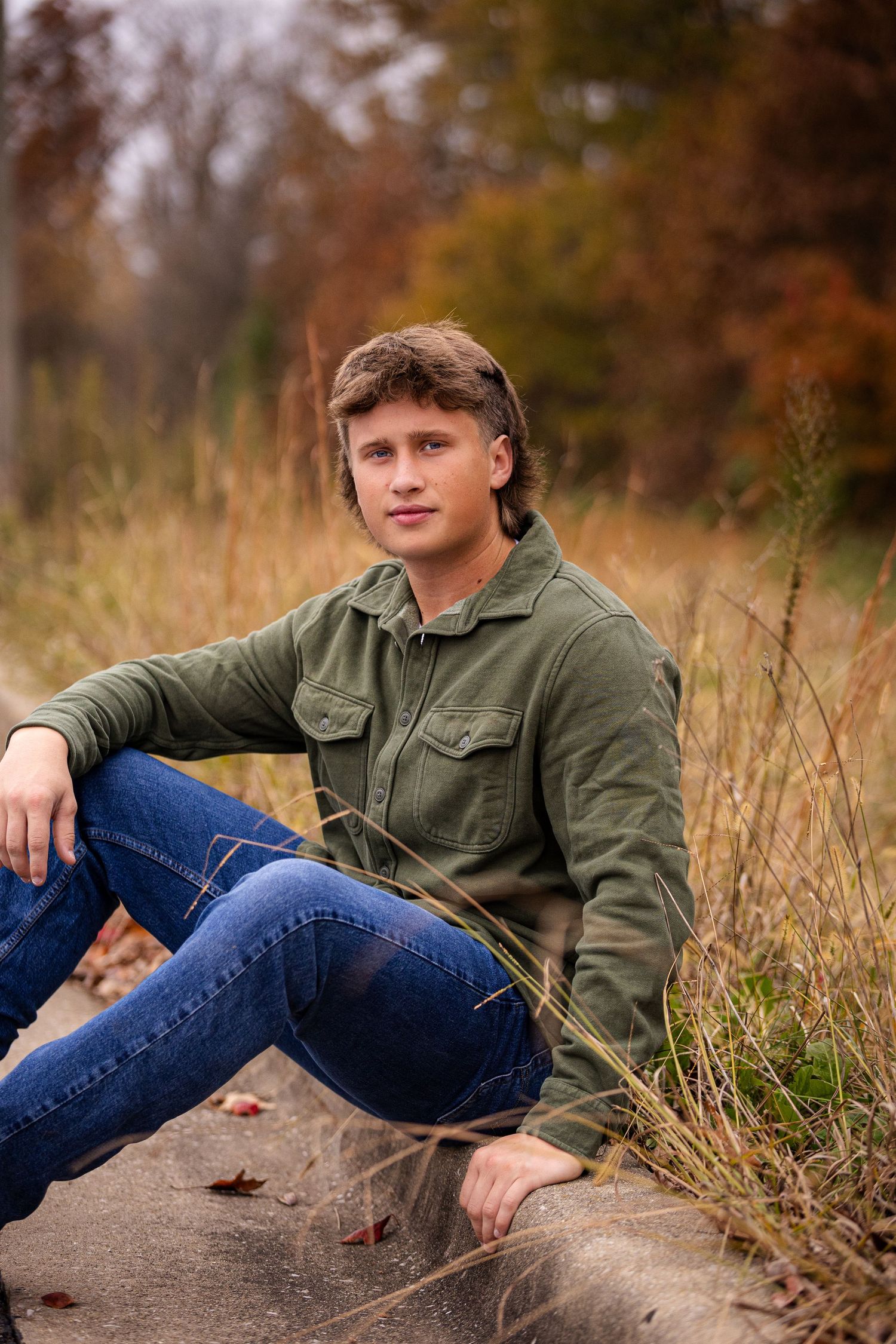 Young man in olive jacket sitting casually outdoors among autumn foliage.