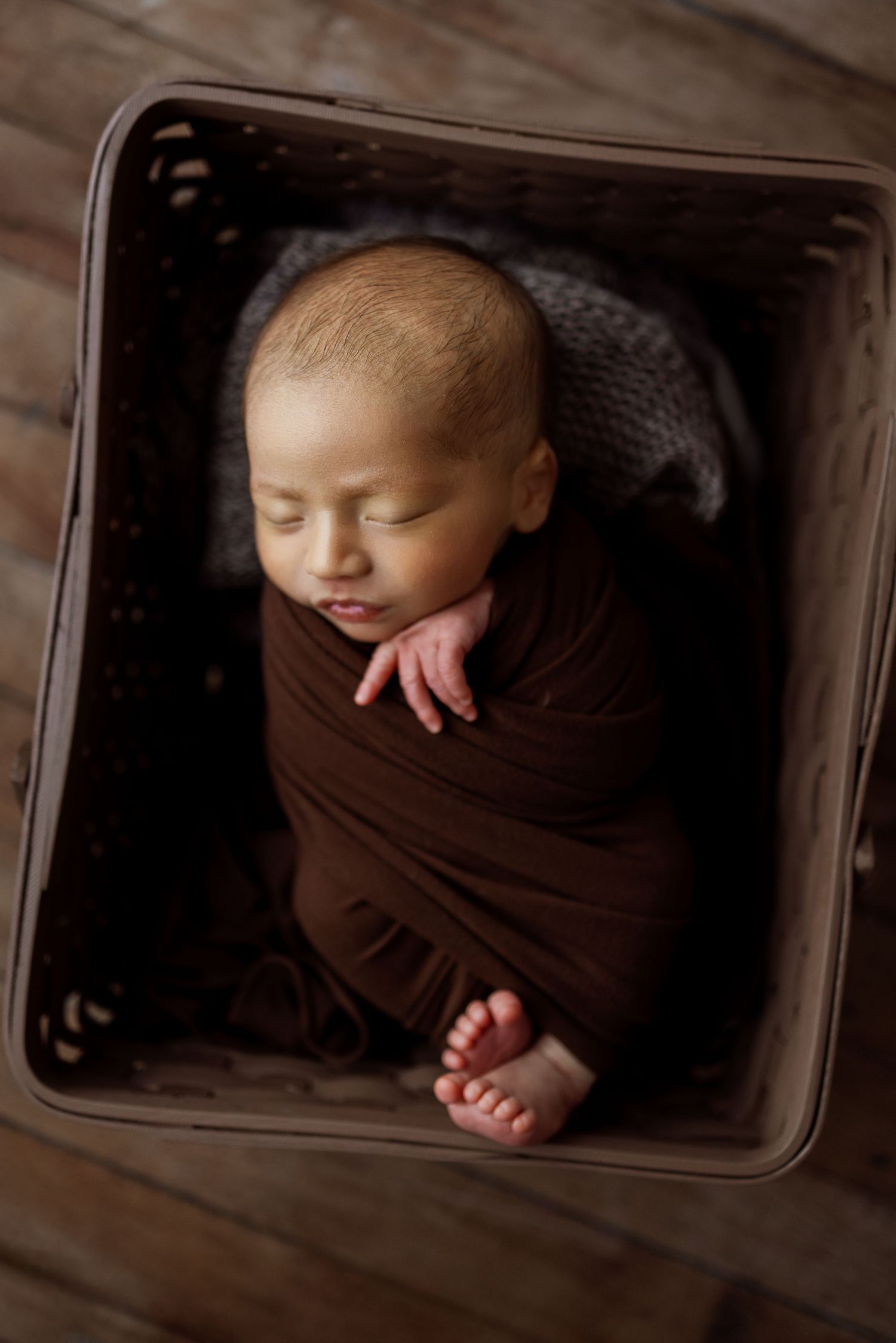 Newborn baby wrapped in brown fabric lying in rustic wooden crate in artistic portrait series.