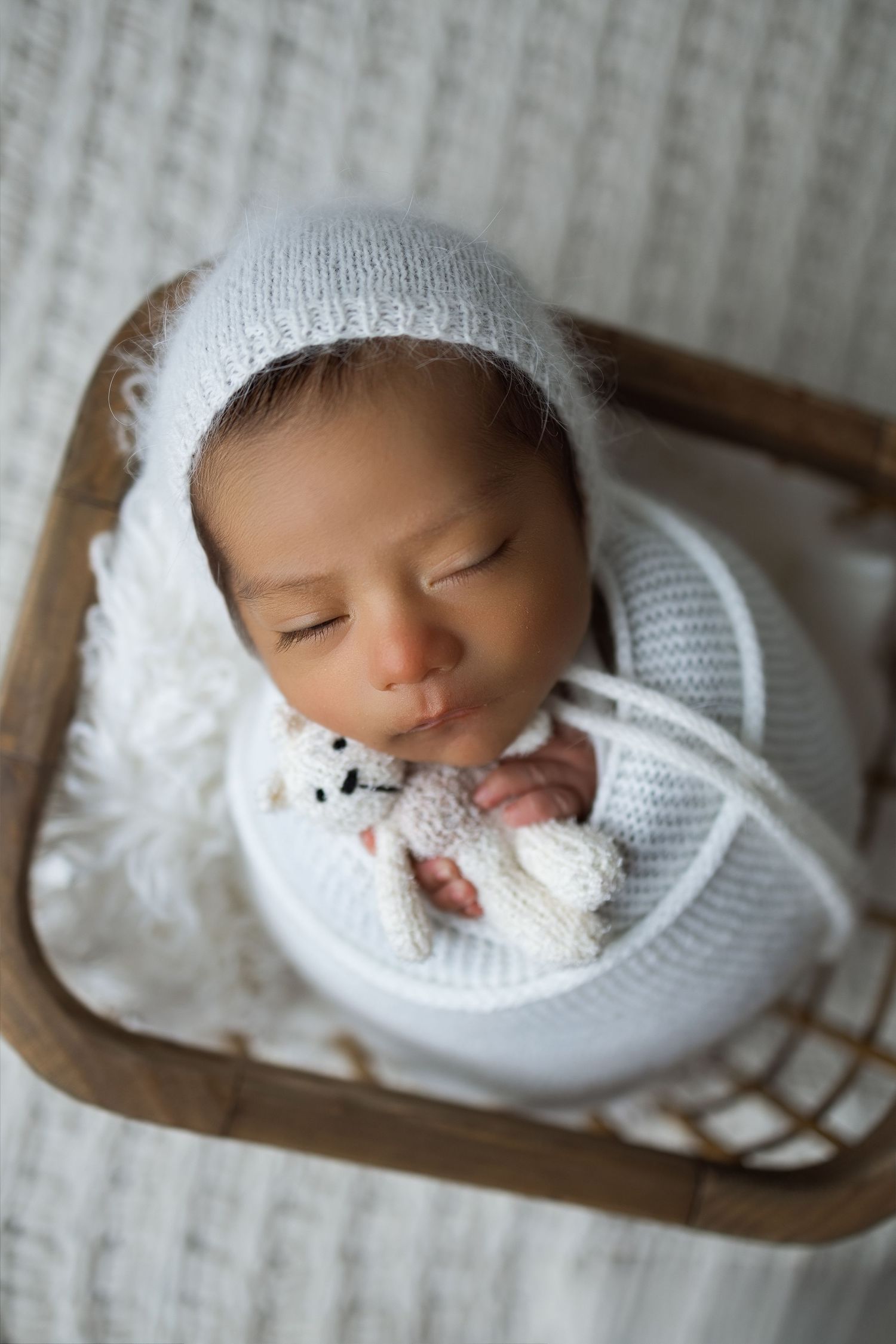Sweet newborn baby sleeping peacefully in white knit wrap on wooden prop against neutral background.