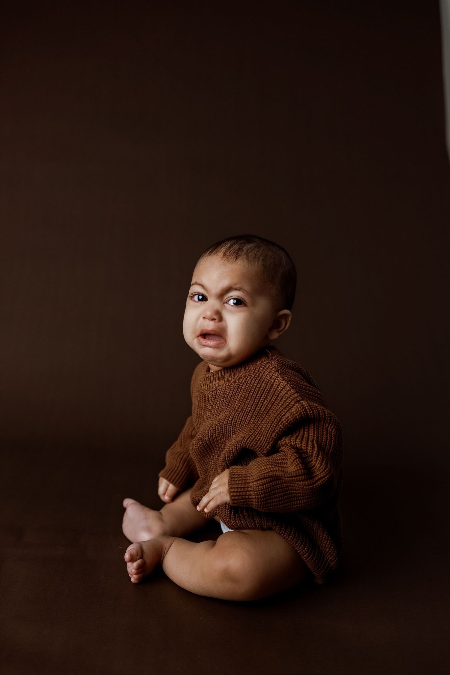 Baby in brown sweater poses against dark background in artistic portrait series.