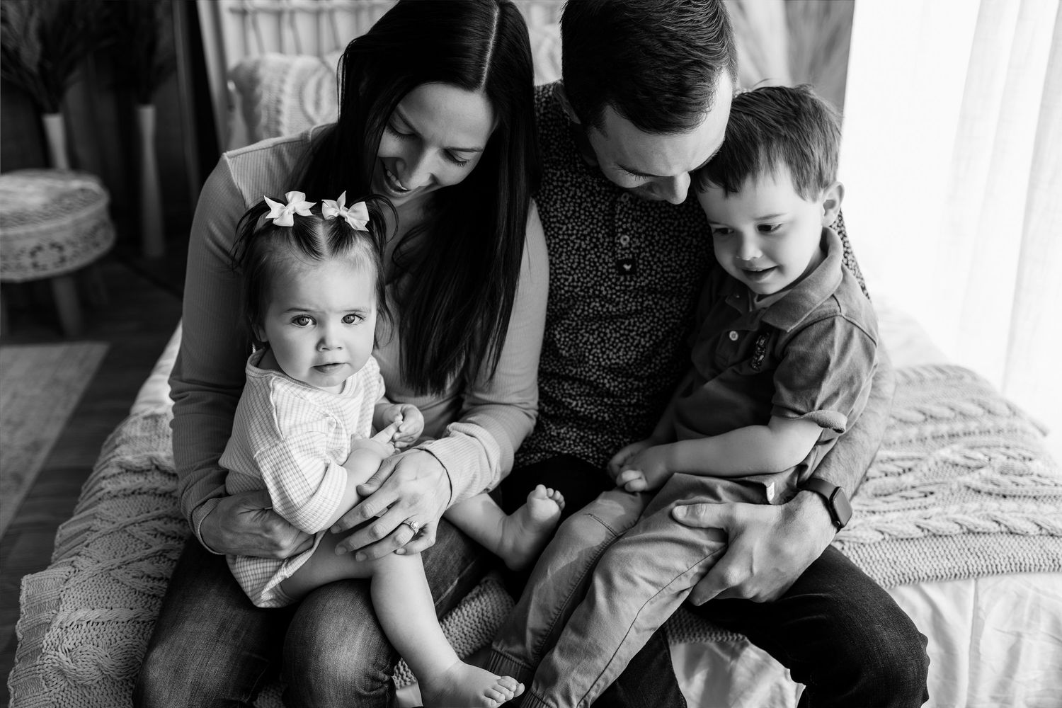 Black and white family portrait of parents sitting on bed with toddler girl and young boy, smiling warmly.