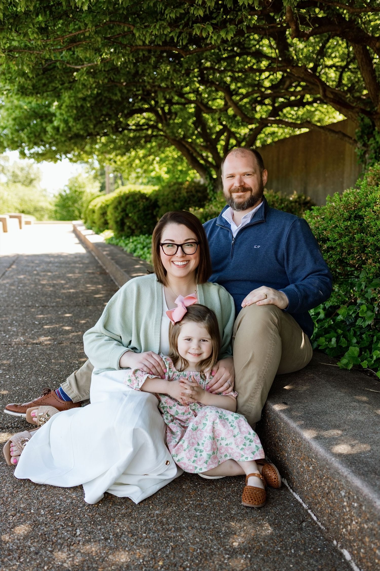 Family of three posing together beneath green trees during an outdoor family photography session
