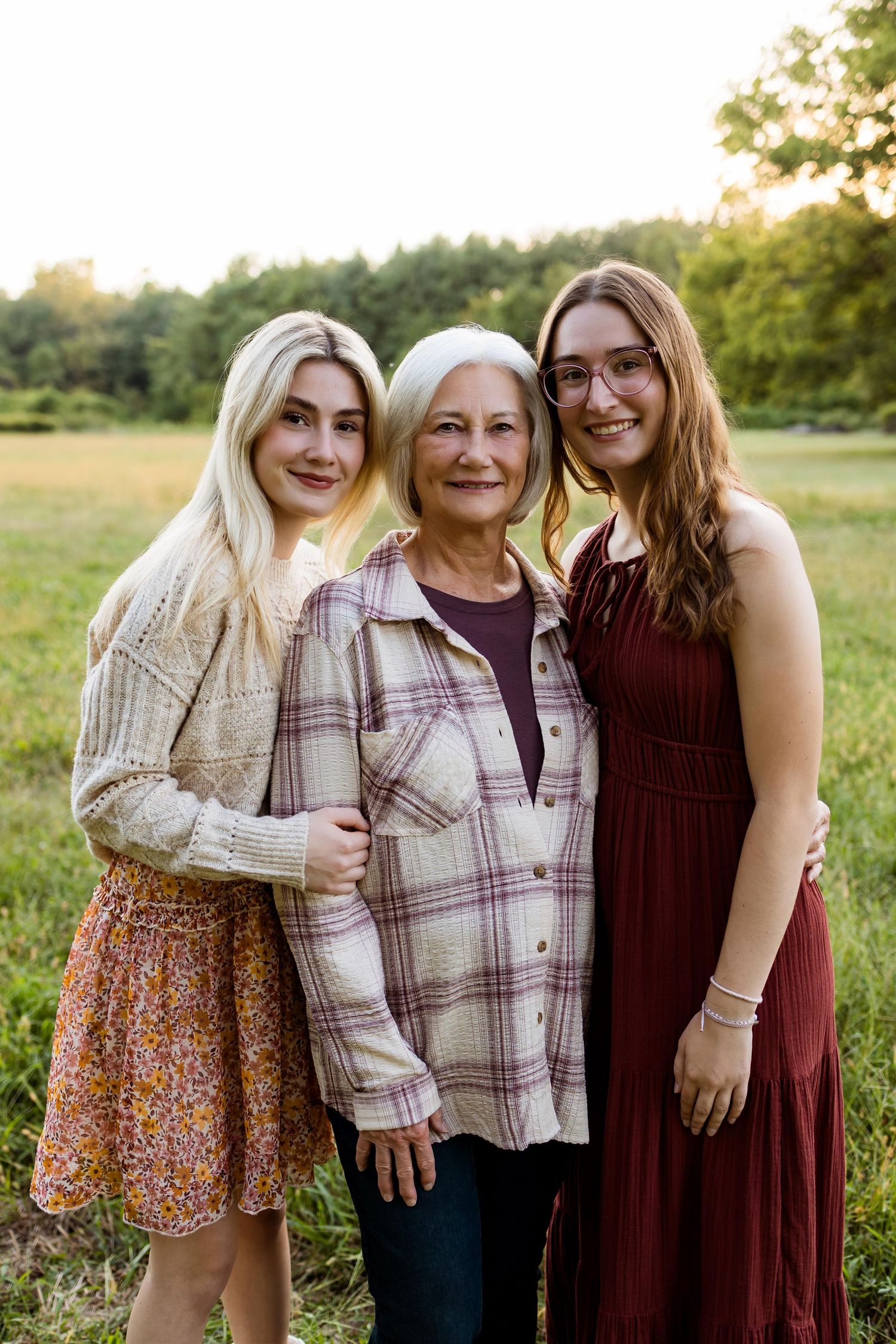 Three generations pose together in a sunny field during golden hour wearing casual fall attire.