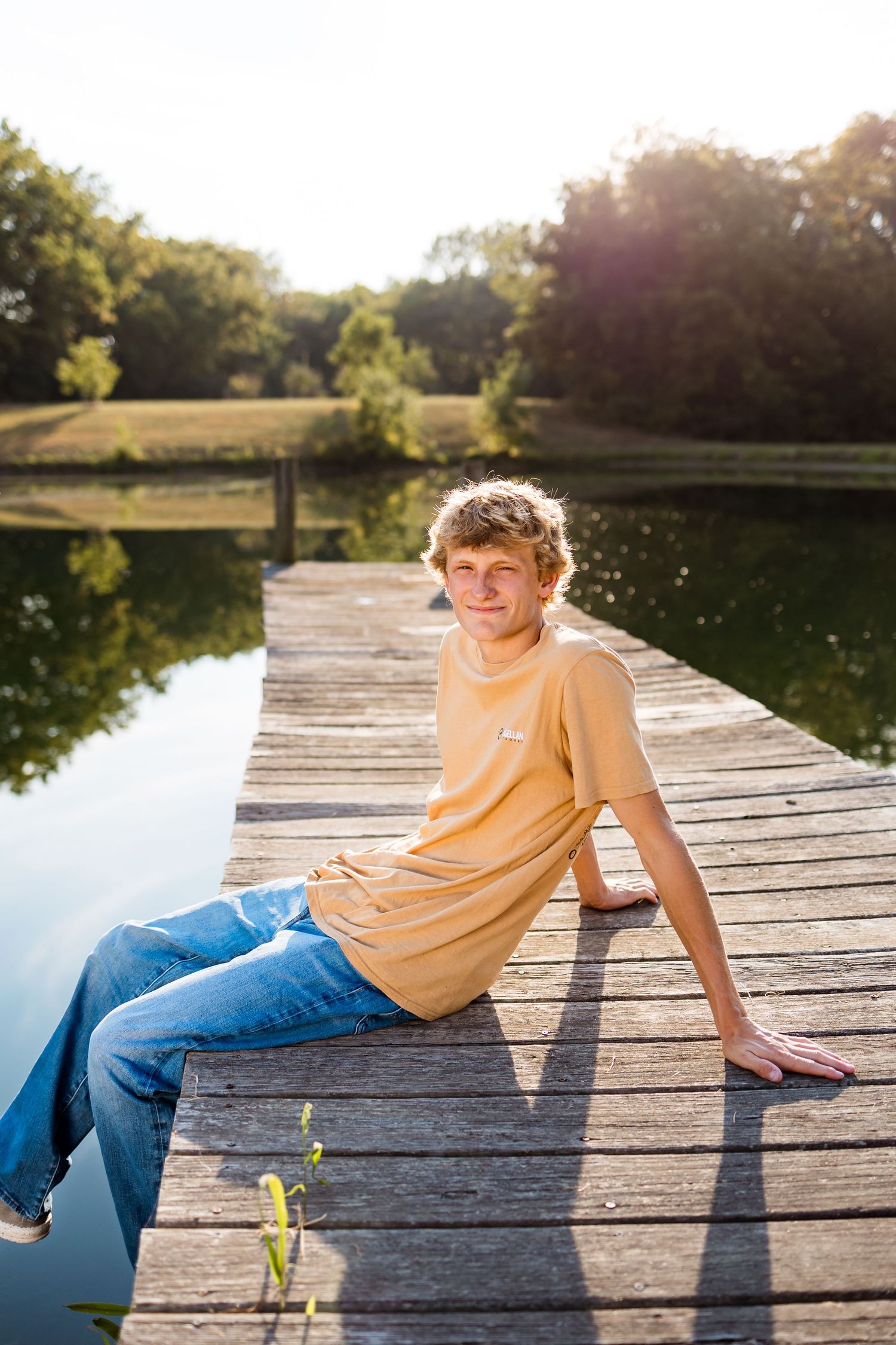 Person in yellow shirt and jeans relaxes on wooden dock by tranquil lake during golden hour.